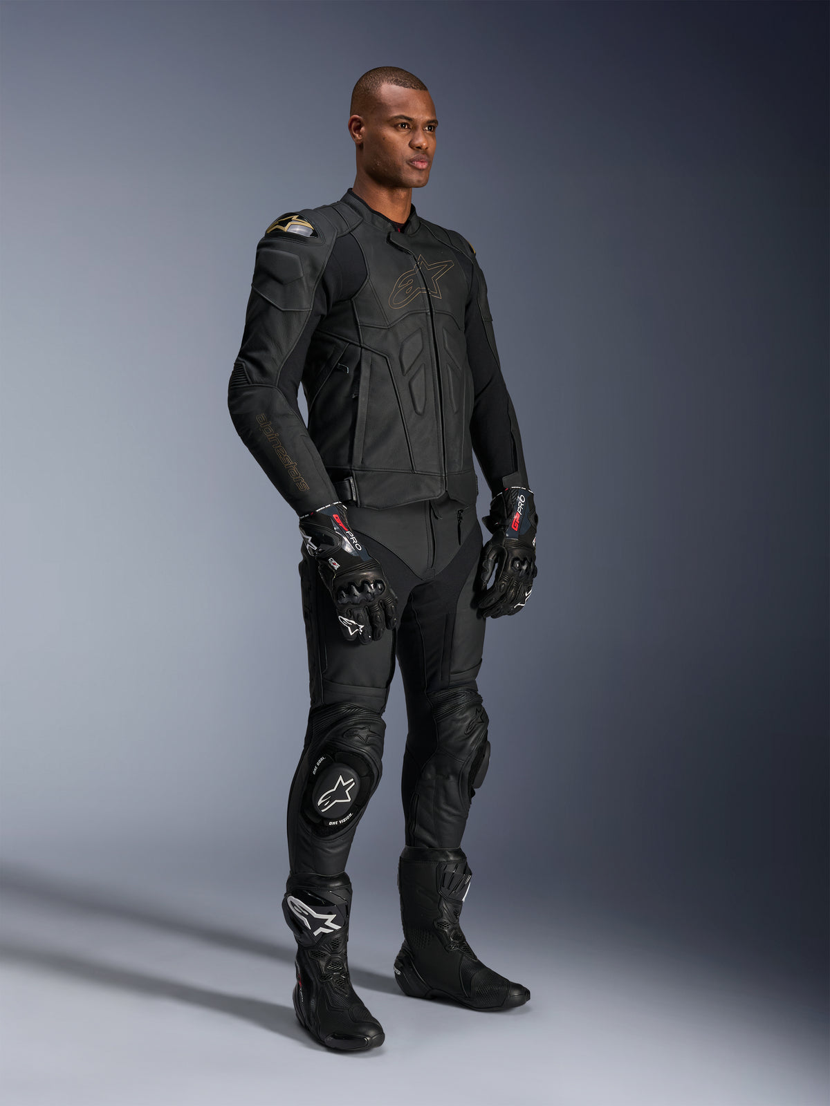 Männliches Model in Alpinestars Missile V3 Lederhose und passender schwarzer Lederjacke mit goldenen Akzenten, schwarze GP Pro Rennhandschuhe und schwarze Rennstiefel, komplette Motorrad-Rennausrüstung für den Straßeneinsatz, Studioaufnahme.