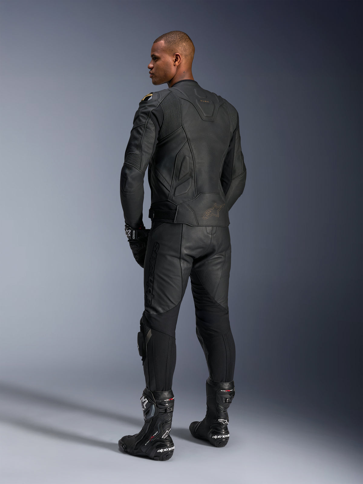 Alpinestars Missile V3 Lederhose und Dusk Lederjacke, Schwarz mit goldfarbenen Akzenten, Rückansicht eines Fahrers, mit geprägtem Branding und Akkordeon-Stretcheinsätzen, getragen mit schwarzen Supertech R Stiefeln und Handschuhen, professionelle Motorsportbekleidung.