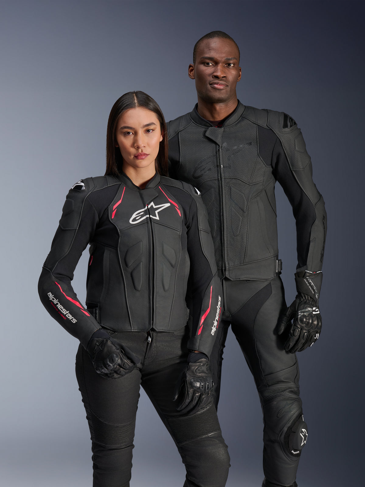 Alpinestars Stella Dusk Lederjacke, Schwarz Diva Pink Weiß, getragen von einem weiblichen Model neben einem männlichen Model in passender schwarzer Lederbekleidung, mit pinkfarbenen und weißen Akzenten, weißem Stern-Logo auf der Brust und schwarzen Motorradhandschuhen