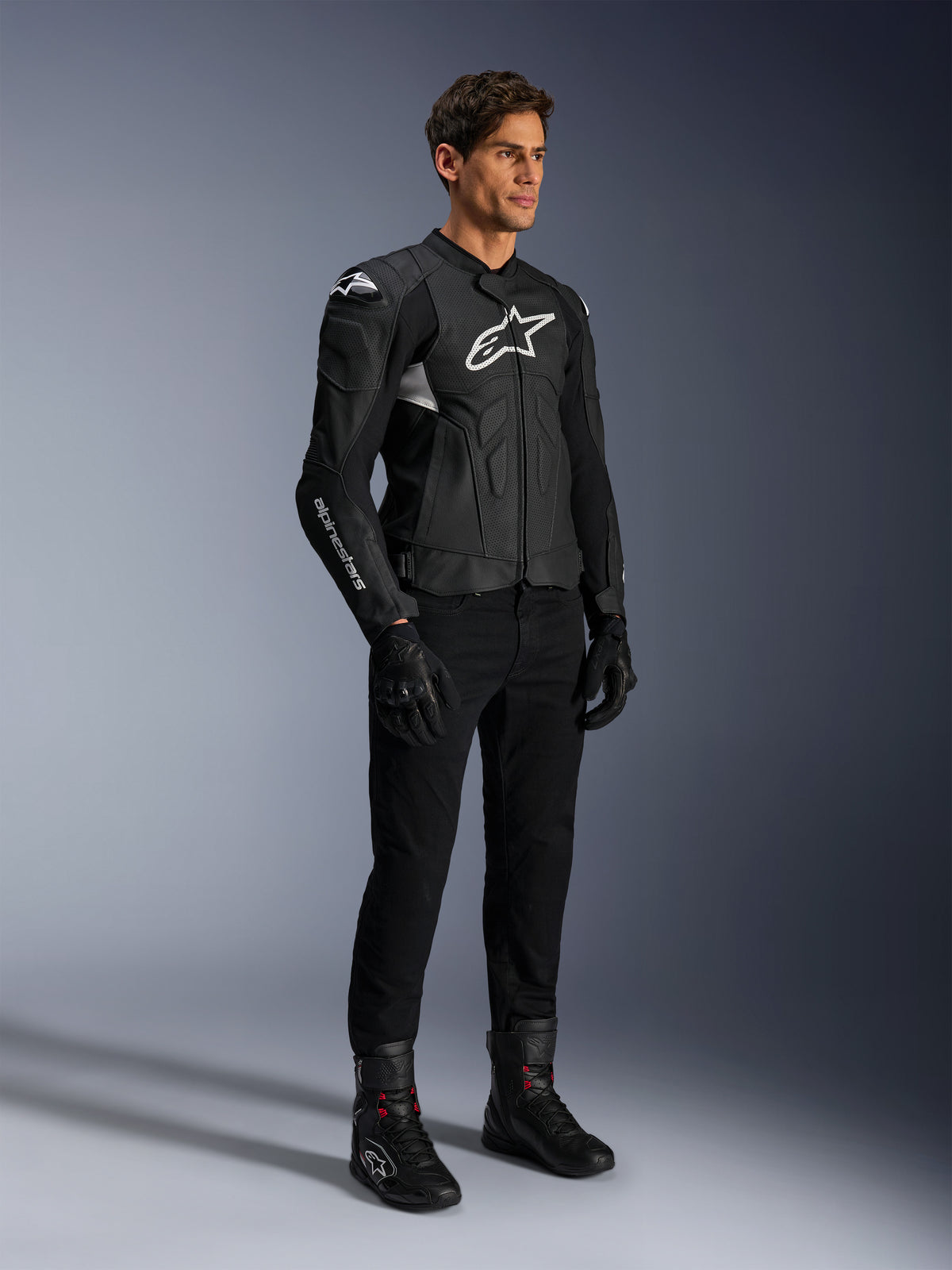 Alpinestars Dusk Airflow Lederjacke, Schwarz und Weiß, präsentiert mit schwarzer Motorradhose, Handschuhen und Stiefeln, mit perforierten Lederbahnen und weißem Logo-Branding, sportlicher Road-Racing-Stil.