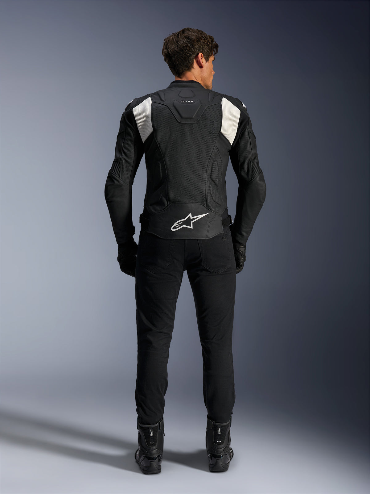 Alpinestars Dusk Airflow Lederjacke, Schwarz und Weiß, Rückansicht eines Fahrers mit schwarzer Hose und Stiefeln, mit perforiertem Leder für Belüftung, weißen Akkordeon-Stretcheinsätzen an den Schultern und weißem Branding am unteren Rücken