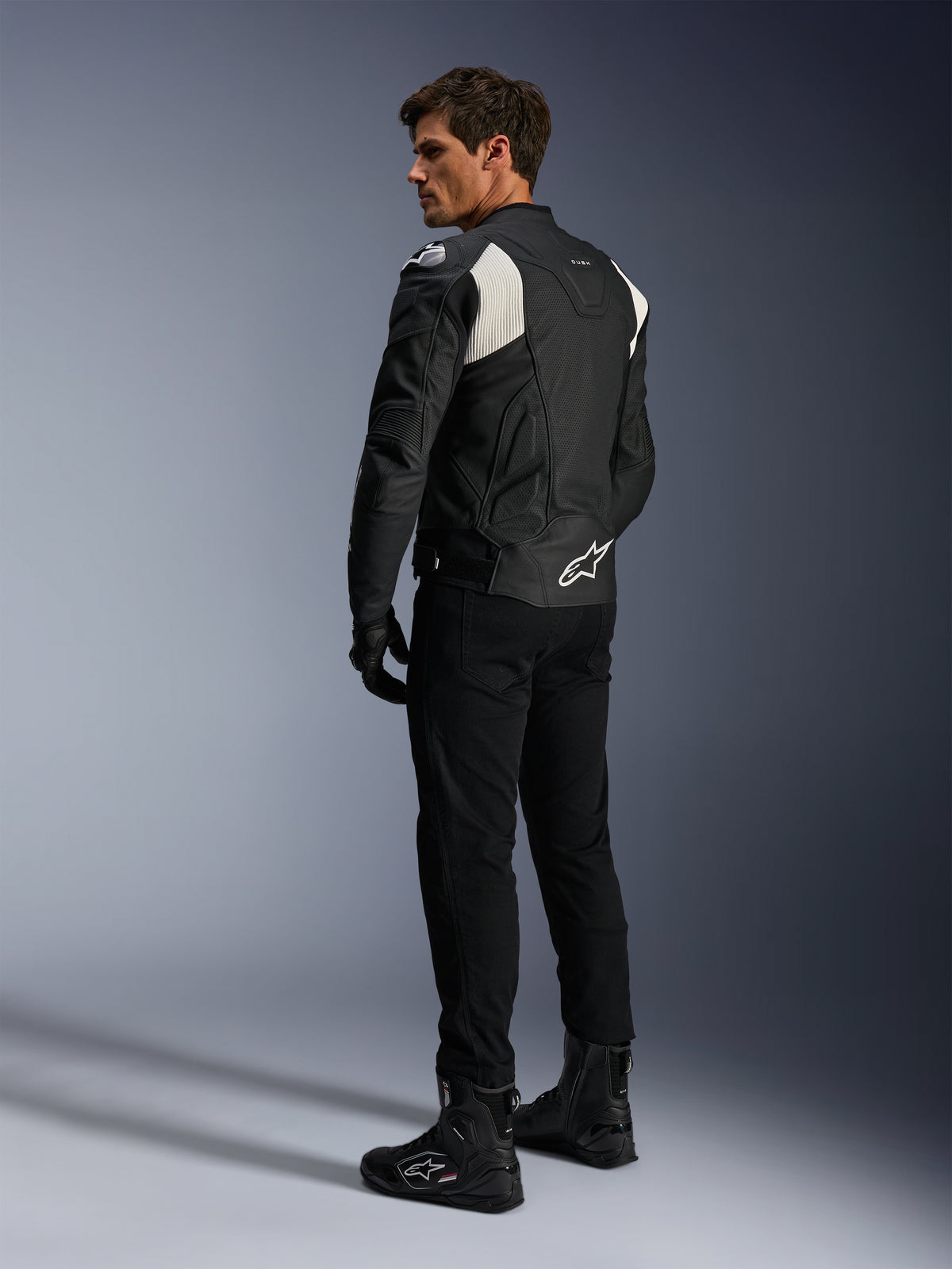 Alpinestars Dusk Airflow Lederjacke, Schwarz und Weiß, Rückansicht eines Models in der perforierten Lederjacke mit passender schwarzer Hose und Stiefeln, mit weißen Akkordeon-Stretcheinsätzen und markantem Logo am unteren Rücken, Sport-Motorradbekleidung