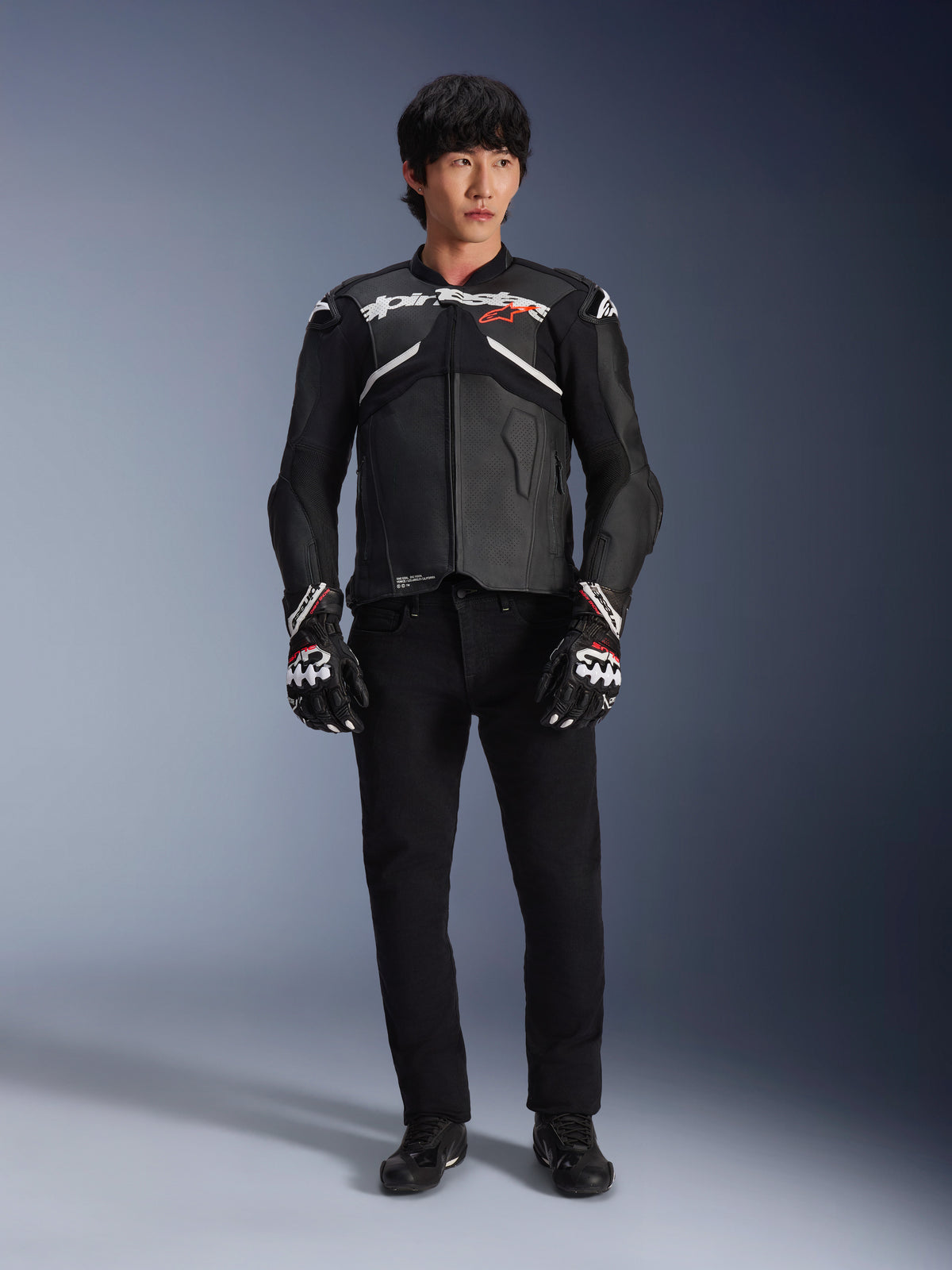 Model trägt die Alpinestars Atem V5 Lederjacke in Schwarz und Weiß, mit weißem Alpinestars-Branding und einem roten Stern-Logo auf der Brust, kombiniert mit einer schwarzen Hose und schwarz-weißen Rennhandschuhen, elegante Performance-Motorradausrüstung