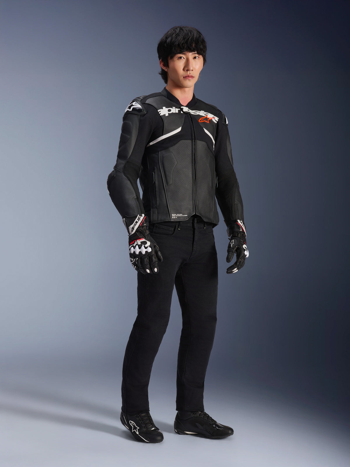Alpinestars Atem V5 Lederjacke, Motorrad-Rennjacke, Schwarz und Weiß, getragen mit schwarzer Hose und passenden Handschuhen, mit weißem Alpinestars-Branding auf der Brust und schützenden Schulterprotektoren.