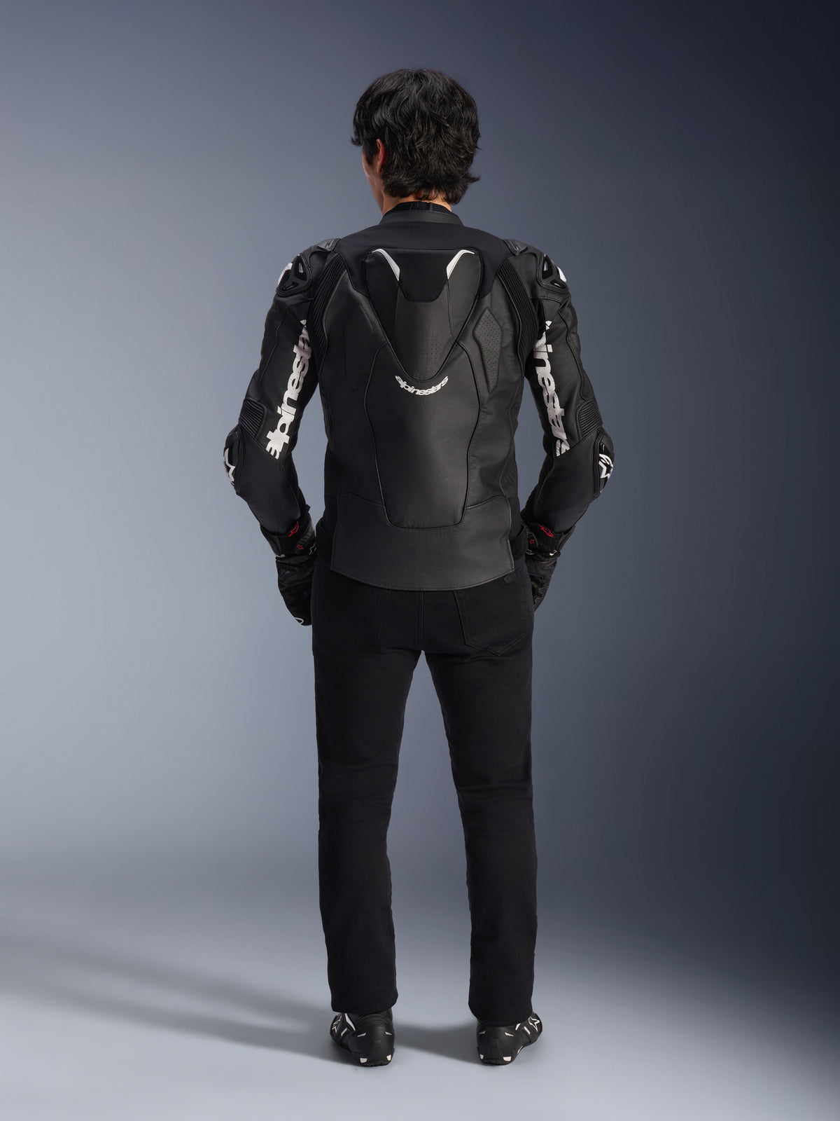 Alpinestars Atem V5 Lederjacke, Motorrad-Rennjacke, Schwarz und Weiß, Rückansicht am Fahrer, mit aerodynamischem Geschwindigkeitsbuckel und weißem Alpinestars-Branding auf Ärmeln und Rücken, technische Lederkonstruktion, kombiniert mit schwarzer Hose und Motorradstiefeln.