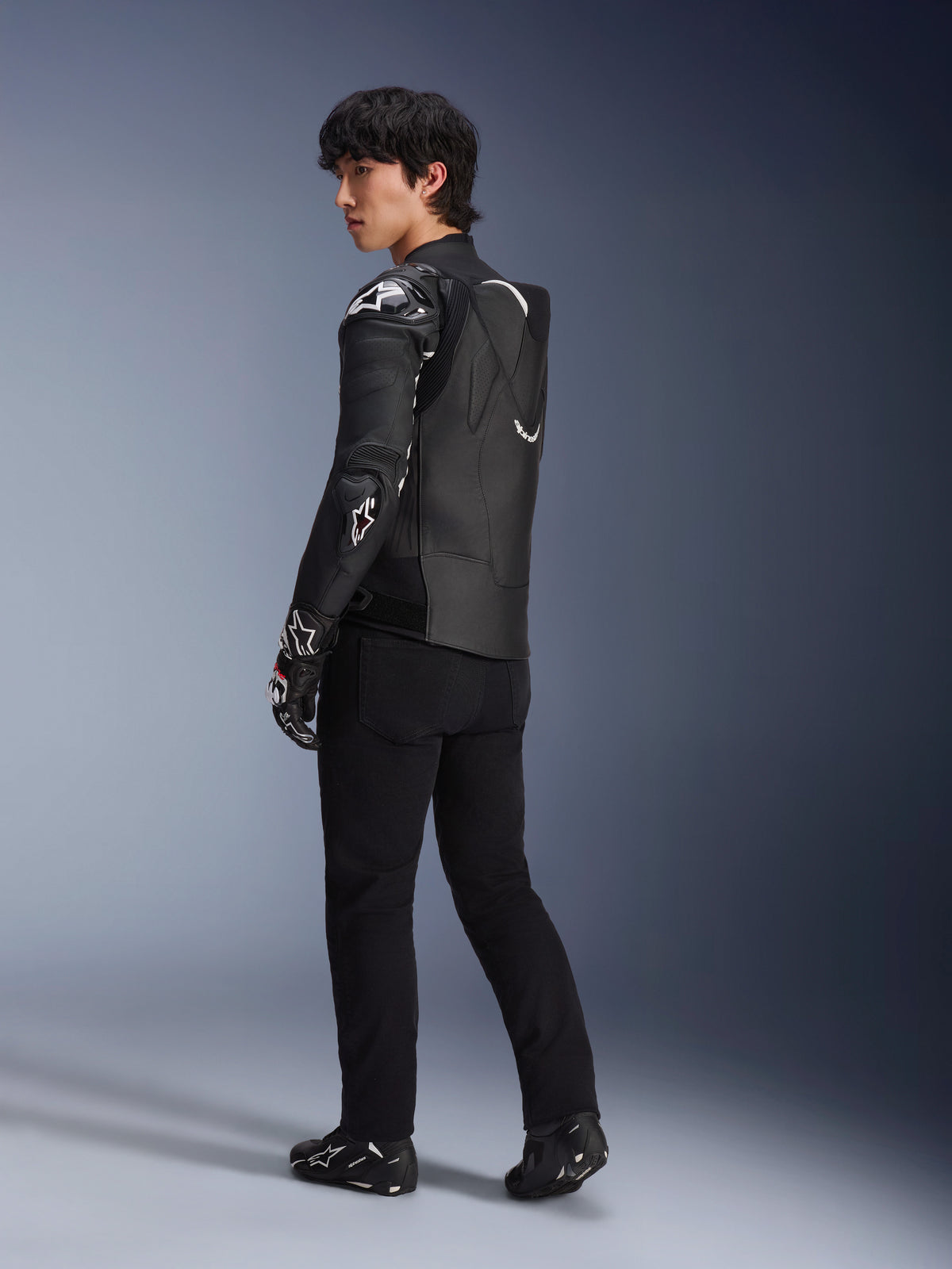 Männliches Model in der Alpinestars Atem V5 Lederjacke in Schwarz und Weiß, Dreiviertel-Rückansicht mit Darstellung des aerodynamischen Speed-Höckers und der DFS RR Protektoren, kombiniert mit schwarzer Hose, Rennhandschuhen und Stiefeln, professionelle Motorrad-Rennbekleidung