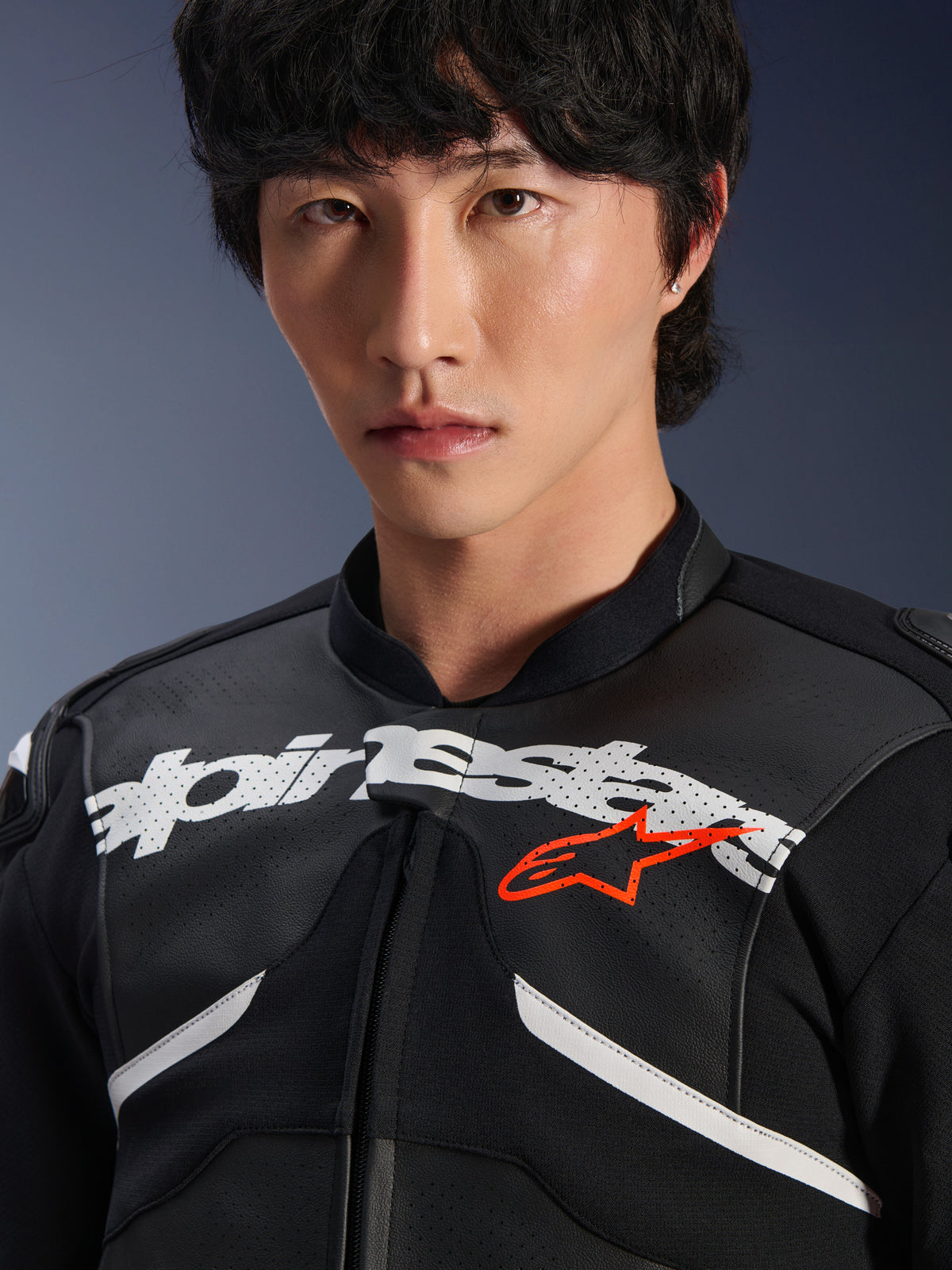 Männliches Model trägt die Alpinestars Atem V5 Lederjacke in Schwarz und Weiß, Nahaufnahme mit dem großen weißen Alpinestars-Logo auf der Brust, rotem Stern-Emblem, perforierter Lederstruktur und technischen Stretch-Einsätzen, Motorsport-Lifestyle-Bekleidung