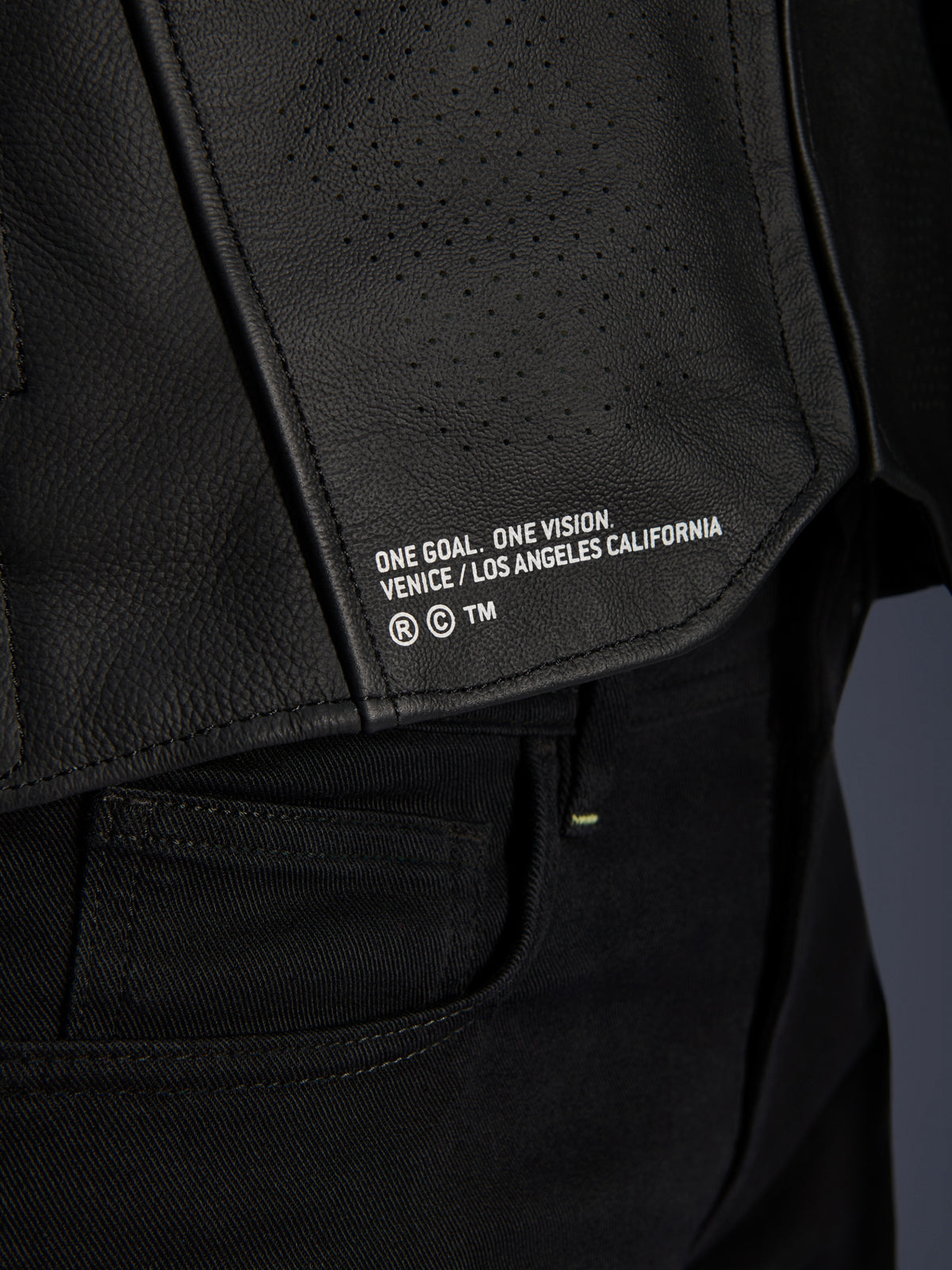 Nahaufnahme der Alpinestars Atem V5 Lederjacke in Schwarz, die den unteren Rückenbereich mit dem „ONE GOAL. ONE VISION. VENICE / LOS ANGELES CALIFORNIA“ Branding und perforierter Lederstruktur zeigt.