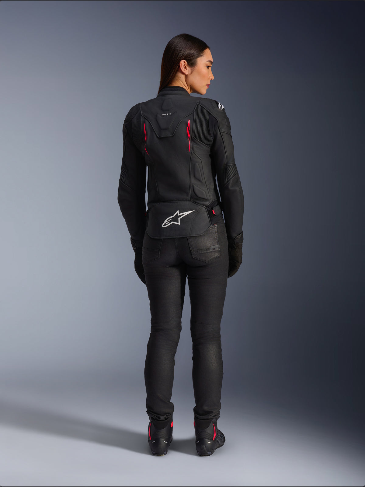 Alpinestars Stella Dusk Lederjacke, Rückansicht, schwarzes Leder mit Akzenten in Diva Pink und Weiß, weißes Alpinestars Logo am unteren Rücken und pinke Belüftungsdetails, getragen mit schwarzer Motorradhose und Stiefeln, hochwertige Sport-Motorradausrüstung für Damen