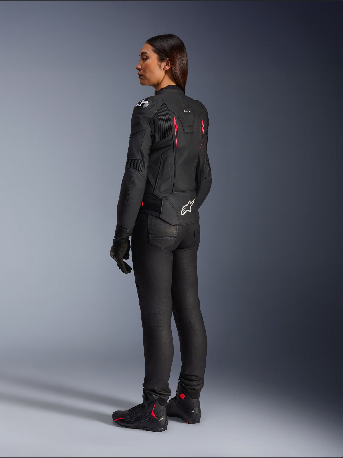 Stella Dusk Damen-Lederjacke, Schwarz Diva Pink Weiß, Dreiviertel-Rückansicht an einer Fahrerin, kombiniert mit schwarzer Motorradhose und schwarzen Stiefeln, hochwertige Lederkonstruktion mit pinken Akzenten und weißem Alpinestars-Logo am unteren Rücken