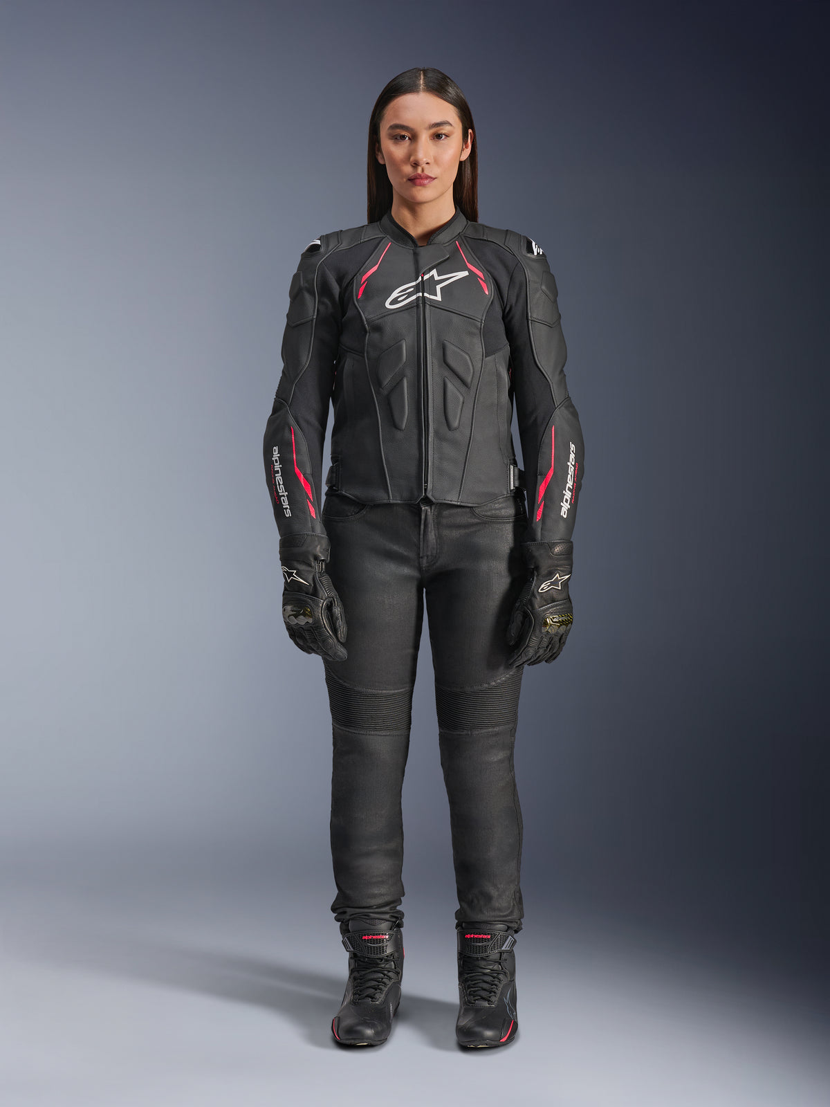 Eine Frau trägt die Alpinestars Stella Dusk Lederjacke in Black Diva Pink White, mit einem weißen Brustlogo und pinkfarbenen Akzenten an den Schultern und Ärmeln, kombiniert mit einer schwarzen Motorradhose, Handschuhen und Stiefeln.