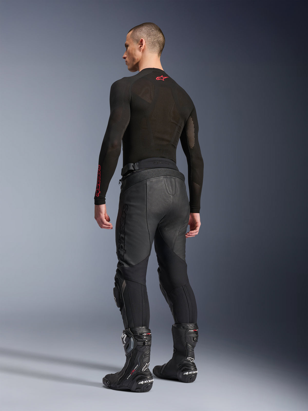 Alpinestars Missile V3 Lederhose, Schwarz, Rückansicht am Modell, getragen mit einem schwarzen langärmeligen Base-Layer und schwarzen Motorrad-Rennstiefeln, mit Akkordeon-Leder-Stretcheinsätzen und vorgeformtem Design für eine ergonomische Passform.