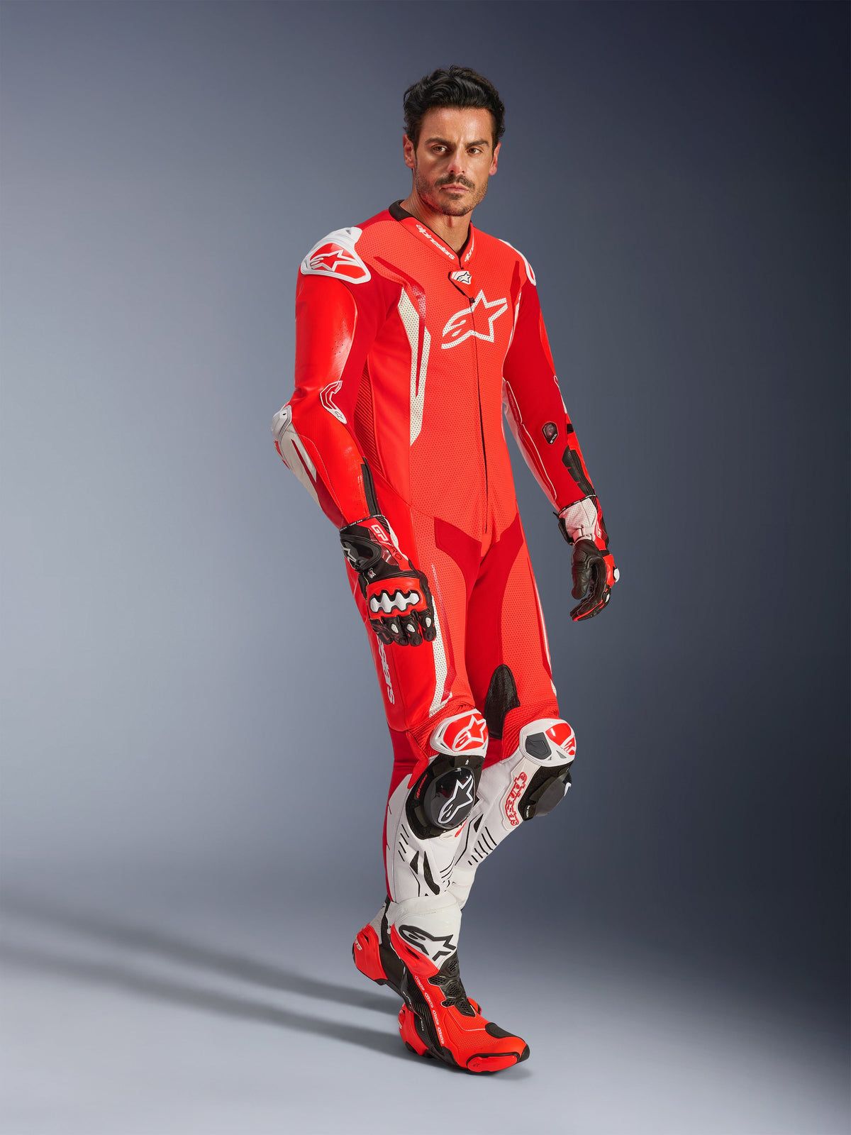 Mann in Alpinestars GP Tech V5 einteiliger Lederkombi, Red Fluo, Hellrot und Weiß, kombiniert mit Rennhandschuhen und Stiefeln, professionelle Motorrad-Rennausrüstung mit aerodynamischem Höcker und integriertem Schutz.