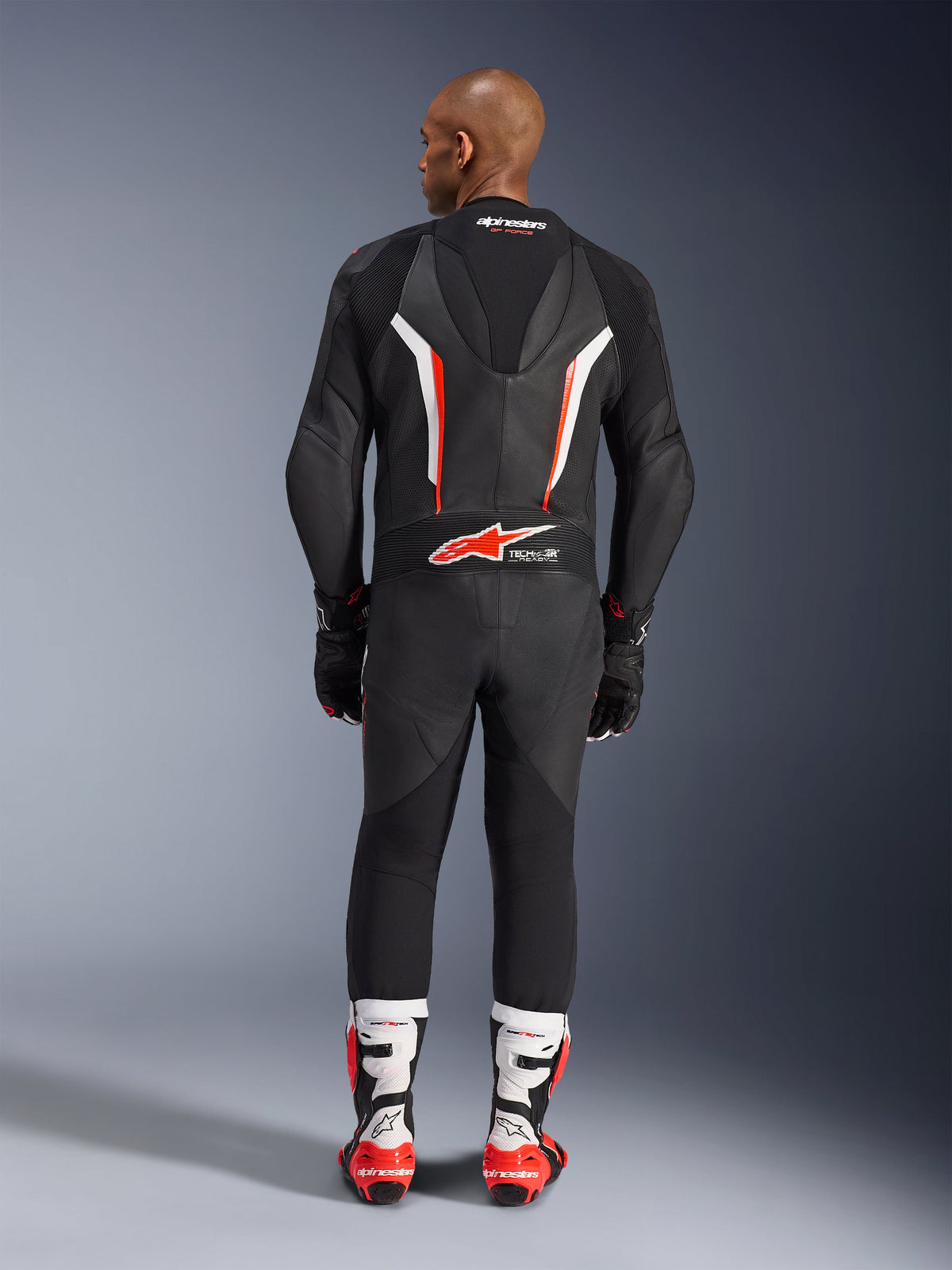 Rückansicht eines Models im Alpinestars GP Force V2 1-Teiler Lederanzug in Schwarz-Weiß-Rot-Fluo, die den aerodynamischen Geschwindigkeitsbuckel und das Tech-Air-Ready-Branding hervorhebt, kombiniert mit weiß-roten Profi-Rennstiefeln und schwarzen Handschuhen.