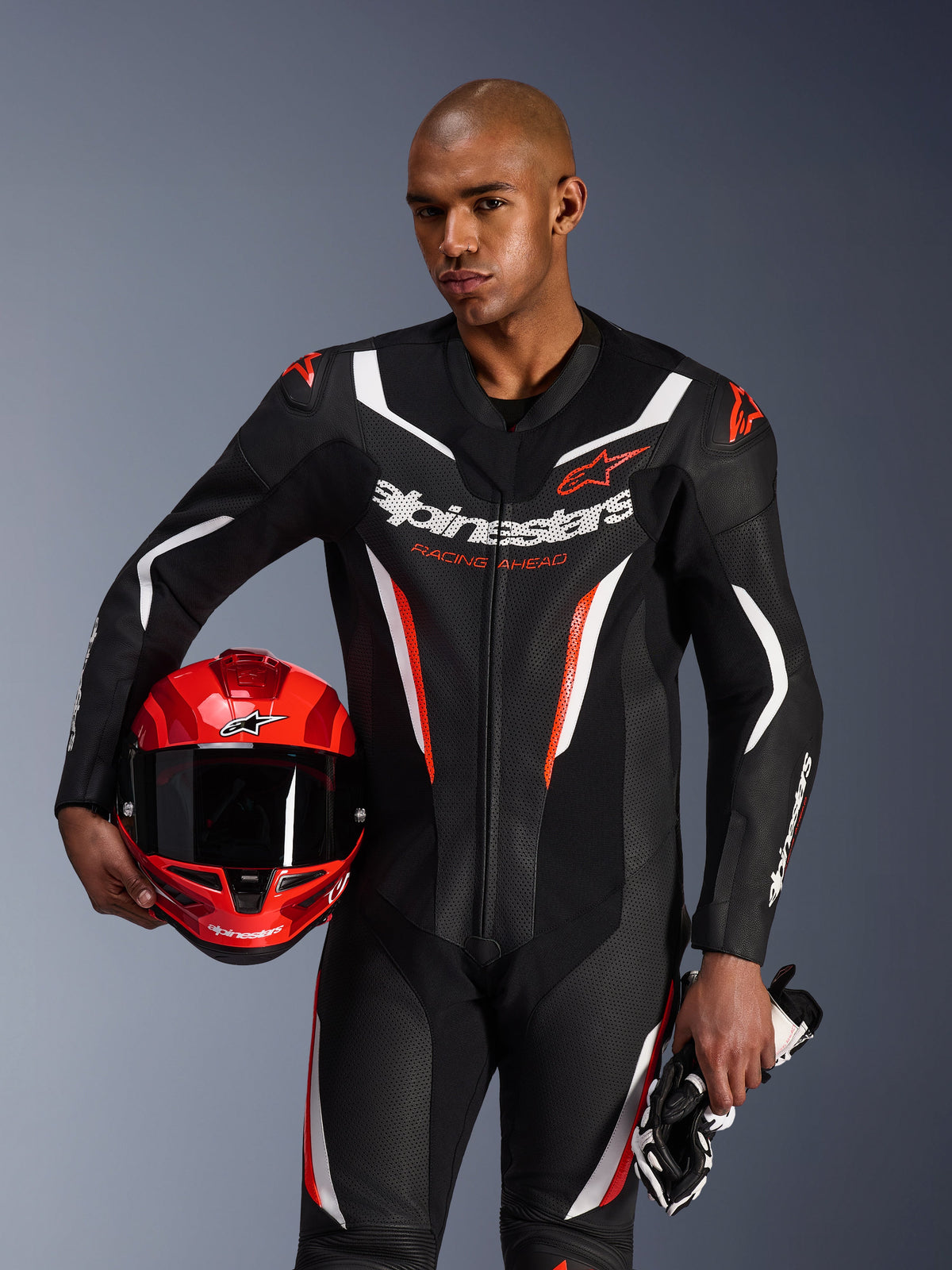 Alpinestars GP Force V2 Lederkombi, 1-teiliger Motorradanzug, Schwarz, Weiß und Fluo-Rot, getragen von einem Model, das einen roten Helm und Handschuhe hält, schlankes Renndesign mit perforiertem Leder und markantem Branding