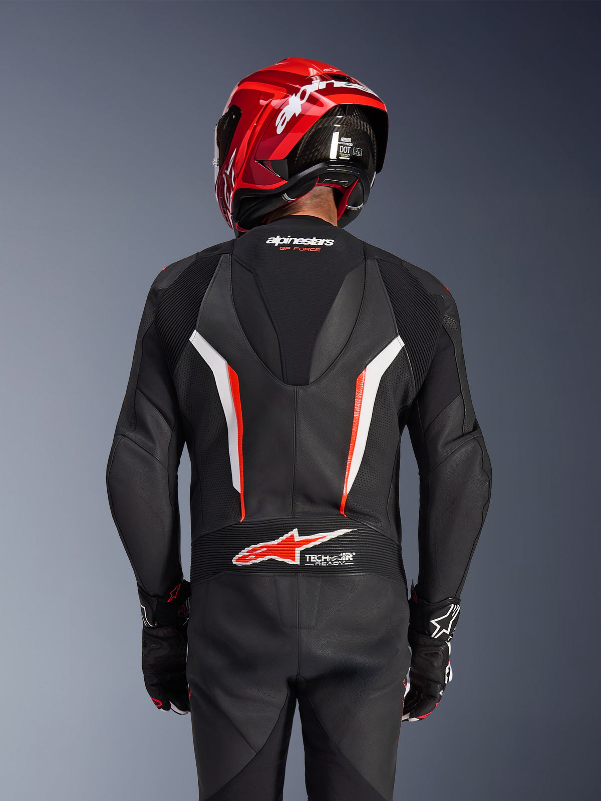 Alpinestars GP Force V2 1-Teiler Lederkombi, Schwarz, Weiß und Rot Fluo, Rückansicht eines Fahrers mit rotem Rennhelm und Handschuhen, mit perforierten Lederbahnen, aerodynamischem Rückenhöcker mit Alpinestars-Branding und Tech-Air Ready-Logo am unteren Rücken, professionelle Motorrad-Rennausrüstung.