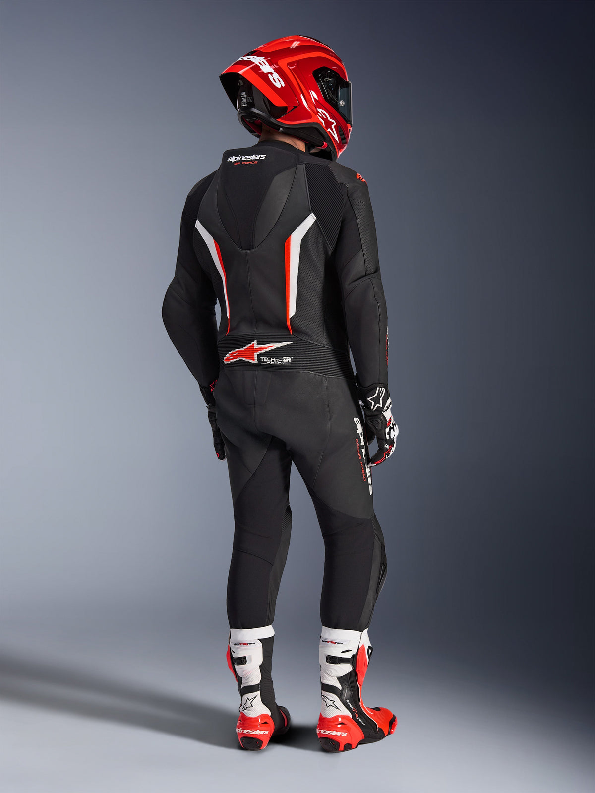 Fahrer in der Rückansicht trägt den Alpinestars GP Force V2 1-Teiler Lederanzug in Schwarz, Weiß und Rot Fluo, mit aerodynamischem Höcker und perforierten Lederpaneelen, kombiniert mit einem roten Alpinestars Helm, Rennhandschuhen und rot-weißen Motorradstiefeln, professionelle Rennstreckenausrüstung.