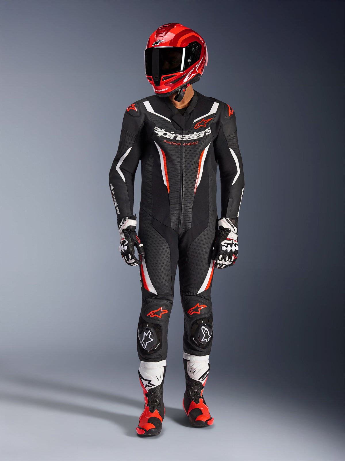 Alpinestars GP Force V2 Lederkombi – 1-Teiler, Schwarz, Weiß und Fluo-Rot, getragen mit einem roten Rennhelm, schwarz-weißen Handschuhen und rot-schwarzen Stiefeln, mit perforierten Lederbahnen und Alpinestars-Branding, professionelle Motorrad-Rennausrüstung