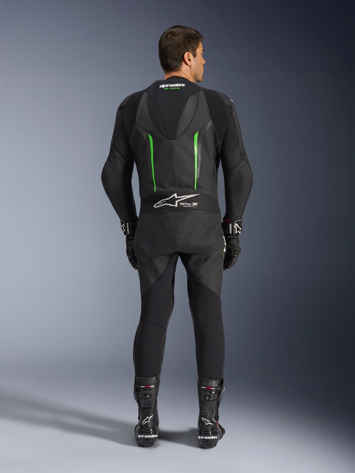 Rear view of the Alpinestars GP Force V2 1-Piece Leather Suit in Black and Green, featuring an aerodynamic speed hump, perforated panels, and Tech-Air Ready branding on the lower back, worn with black racing gloves and boots.