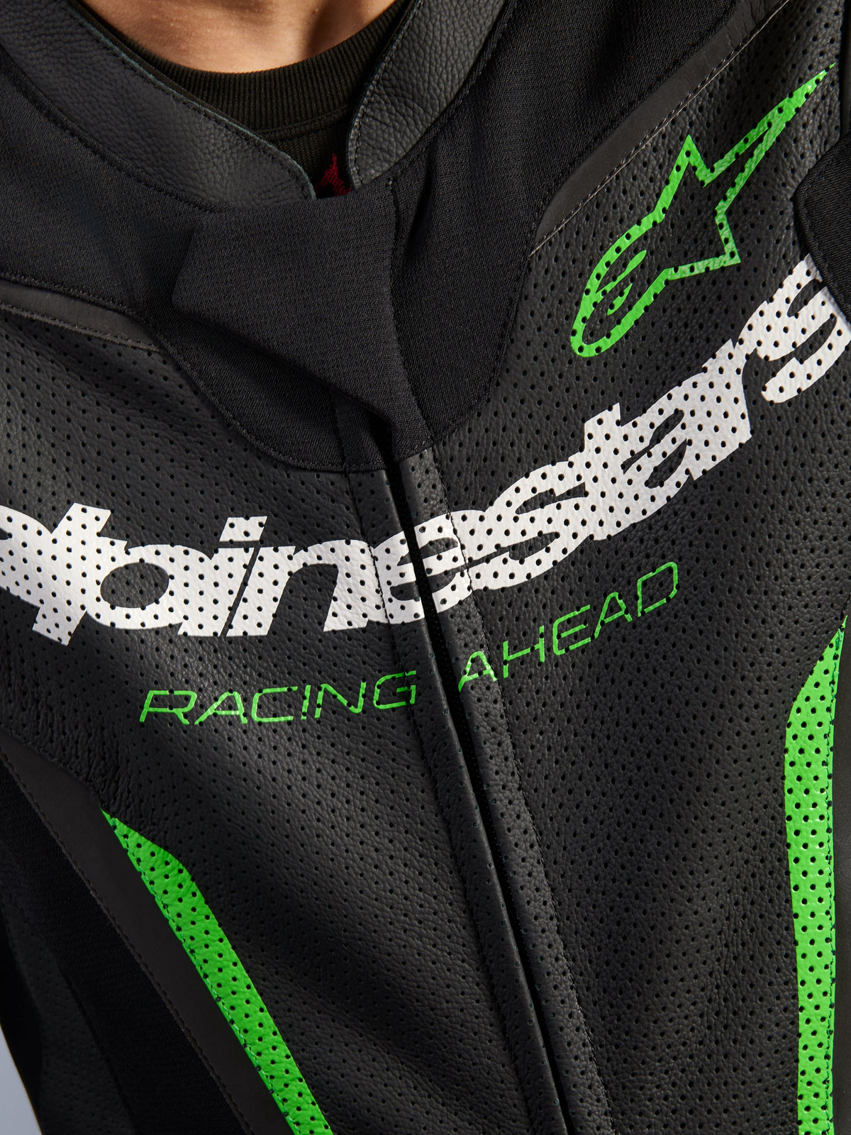Alpinestars GP Force V2 Leather Suit - 1Piece, close-up of chest area, black perforated leather with white Alpinestars branding, green 'RACING AHEAD' text, and green accents