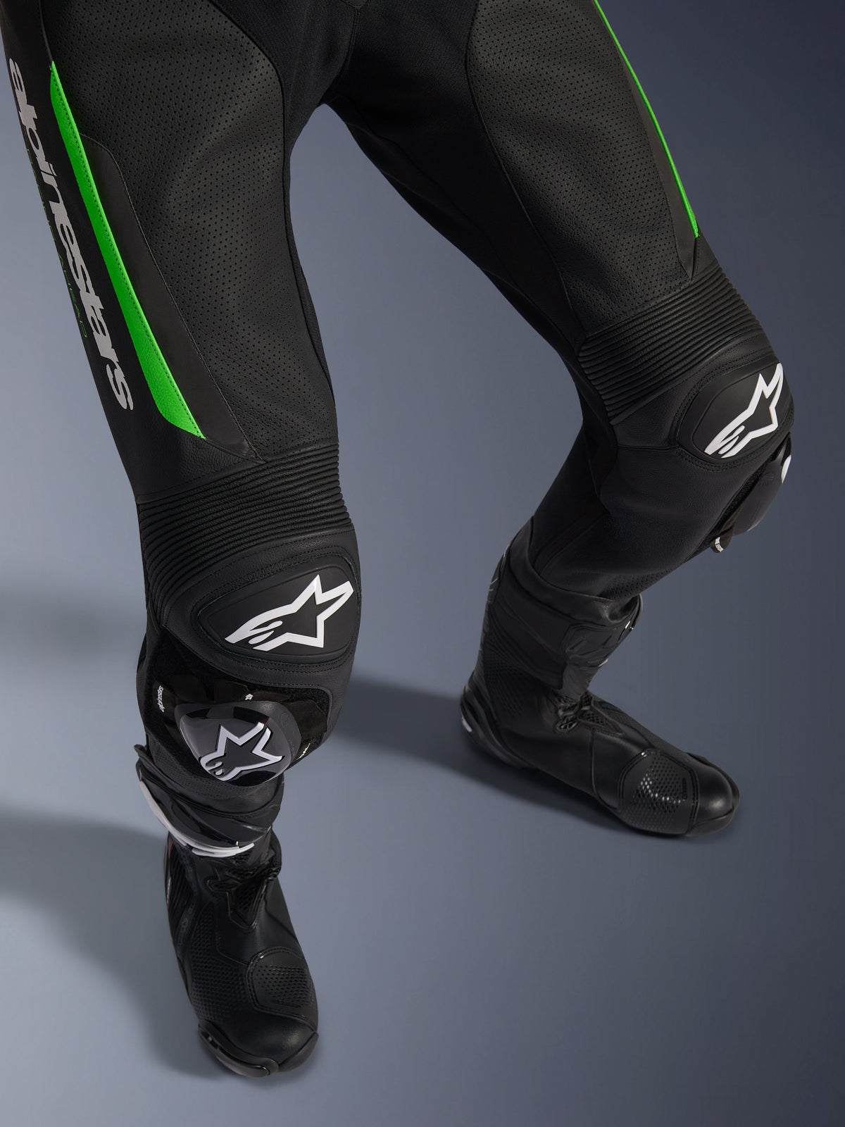 Alpinestars GP Force V2 Leather Suit - 1Piece, close-up of legs in Black and Green, featuring perforated leather construction, accordion stretch panels at the knees, white Alpinestars branding, and integrated knee sliders, worn with black racing boots