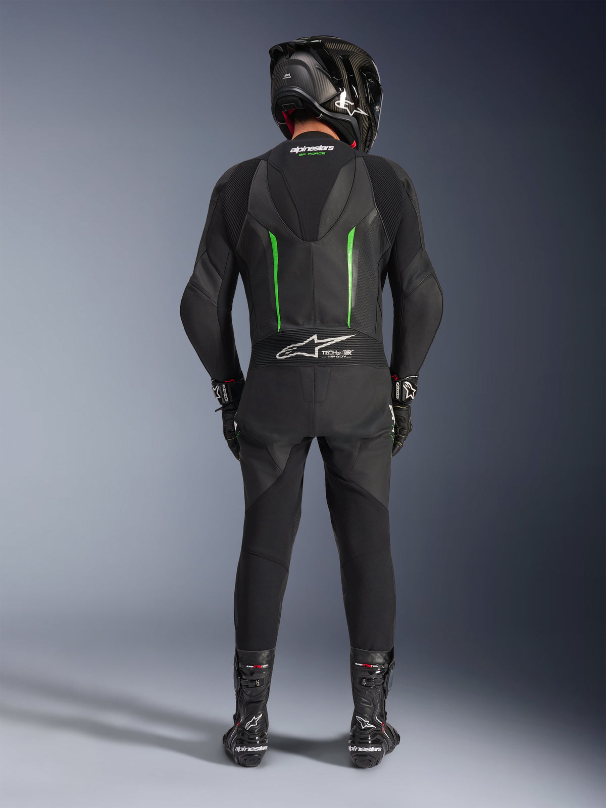 Rider from the back wearing Alpinestars GP Force V2 Leather Suit - 1Piece in Black and Green, paired with a black carbon fiber helmet, black racing gloves, and black boots, featuring an aerodynamic speed hump and Tech-Air Ready branding.