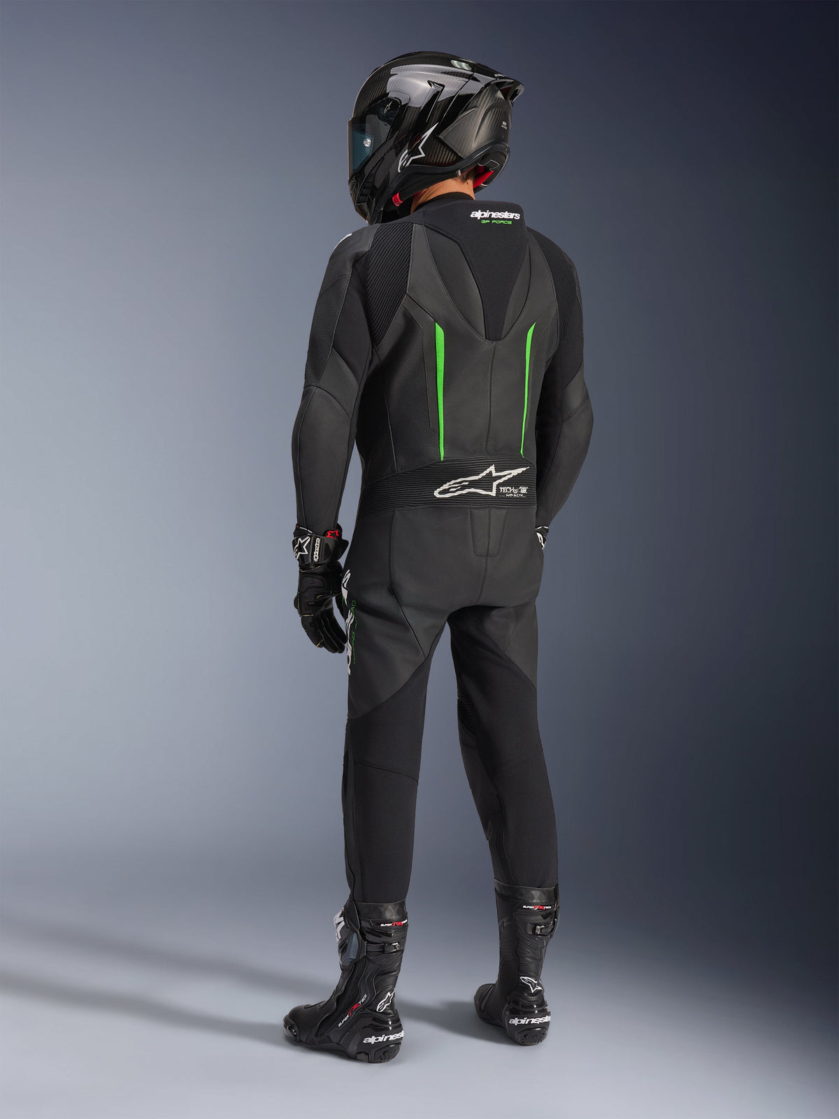 Rider from behind wearing Alpinestars GP Force V2 Leather Suit, 1-Piece, Black with Green accents, aerodynamic speed hump, paired with a black carbon-fiber helmet, black racing boots, and gloves, high-performance motorcycle racing gear