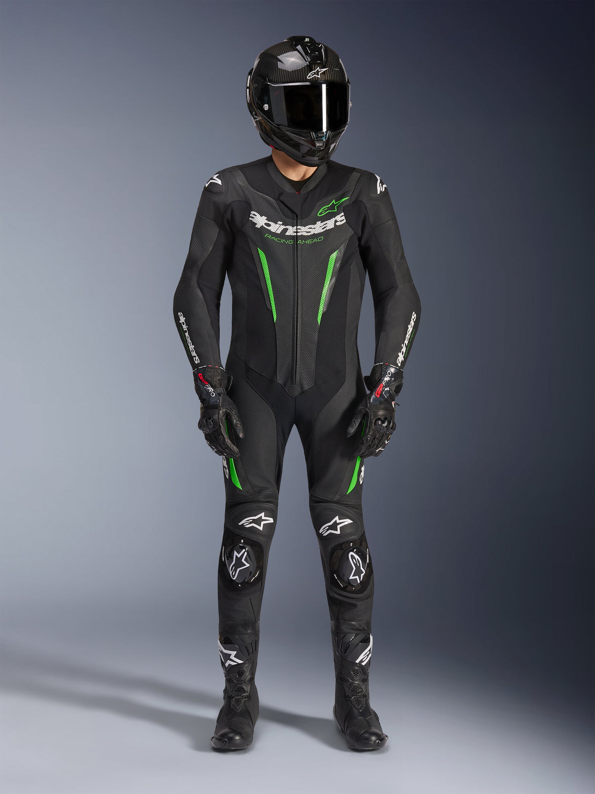Motorcycle racer wearing Alpinestars GP Force V2 Leather Suit - 1Piece in Black and Green, paired with a carbon fiber helmet and racing gloves, featuring perforated leather panels and white Alpinestars logos, professional track and sport riding gear