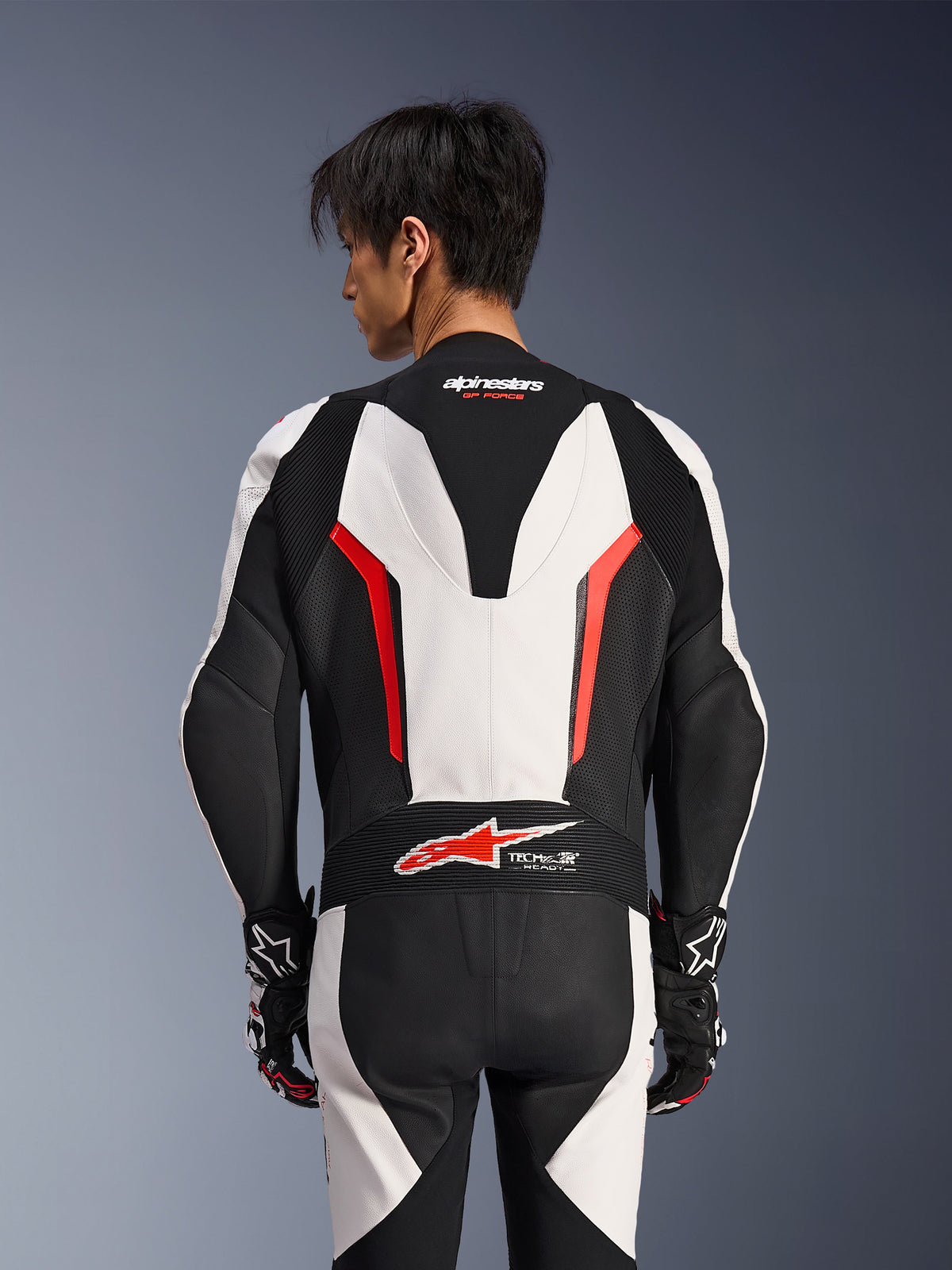 Alpinestars GP Force V2 Lederkombi 1-teilig, Weiß, Schwarz und Rot Fluo, Rückansicht an einem männlichen Model, mit aerodynamischem Höcker, perforierten Lederbahnen und Tech-Air Ready-Logo am unteren Rücken, professionelle Rennausrüstung mit schwarz-weißen Handschuhen