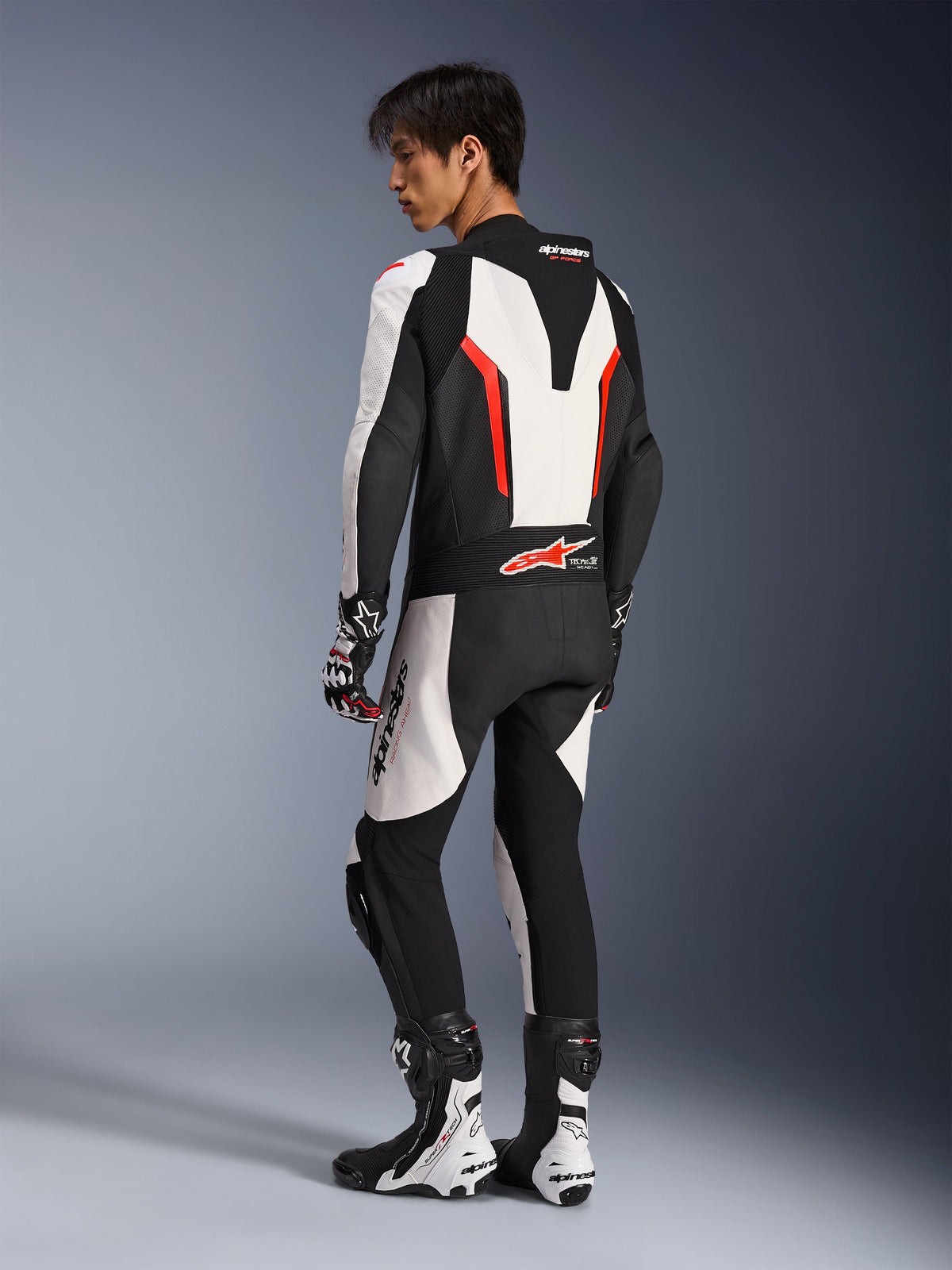 Motorradfahrer im Alpinestars GP Force V2 Lederkombi – Einteiler in Weiß, Schwarz und Rot Fluo, Rückseitenansicht mit aerodynamischem Geschwindigkeitsbuckel und perforierten Lederpaneelen, kombiniert mit passenden Rennstiefeln und Handschuhen, konzipiert für die Rennstrecke und Performance-Fahrten.