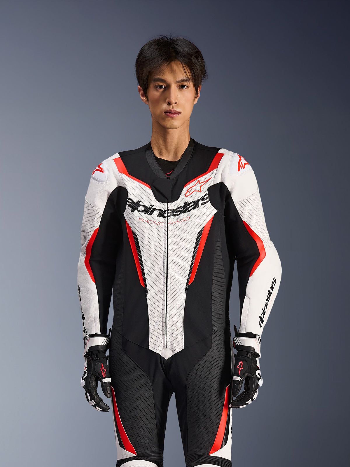 Alpinestars GP Force V2 Lederkombi – 1-teilig, Weiß, Schwarz und Rot Fluo, getragen von einem männlichen Model, mit perforierten Lederpaneelen und markantem Alpinestars-Branding auf der Brust, kombiniert mit schwarz-weißen Rennhandschuhen, entwickelt für die Rennstrecke und Performance-Fahrten
