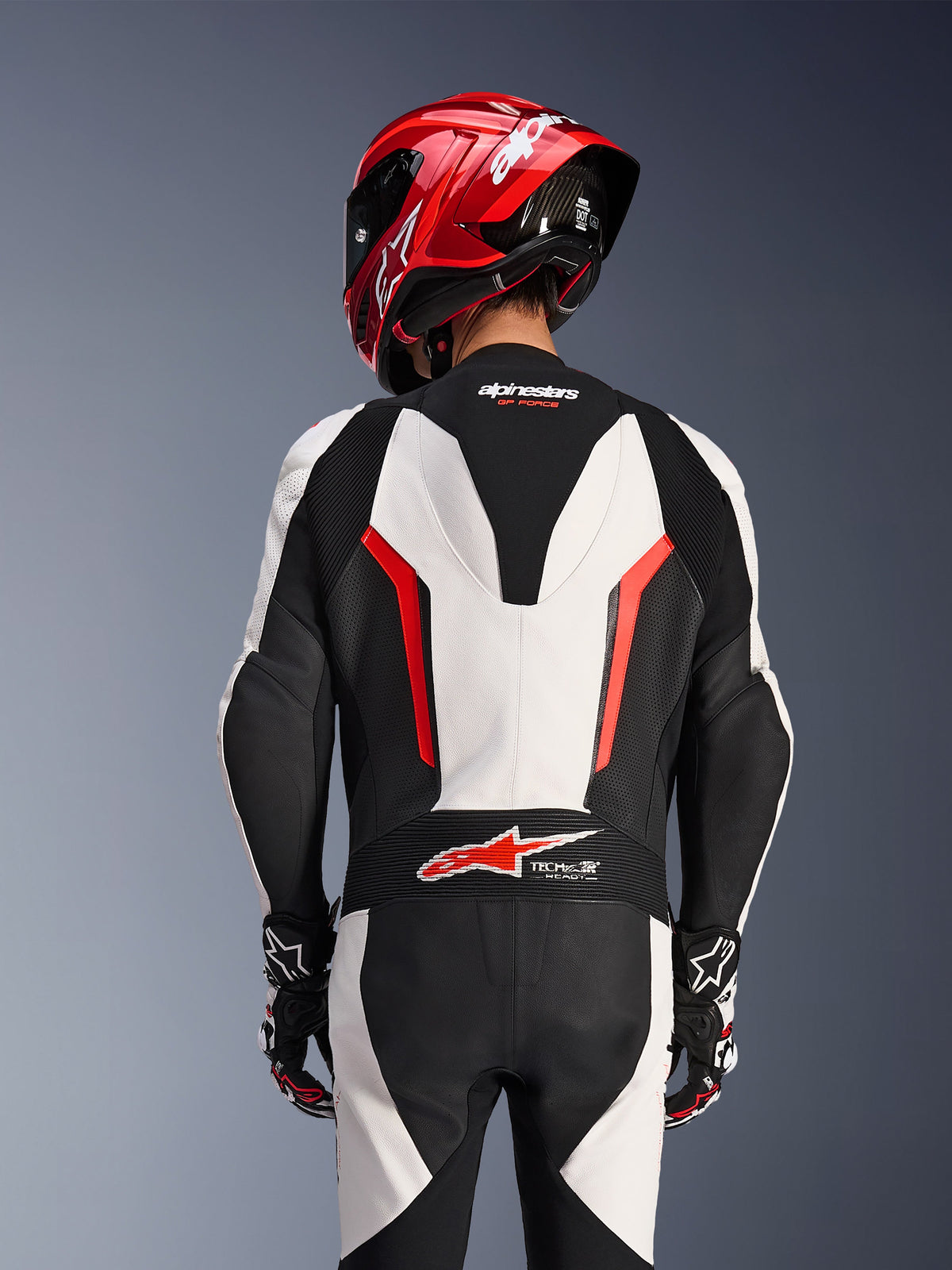 Rückansicht des Alpinestars GP Force V2 1-teiligen Lederkombis in Weiß, Schwarz und Rot Fluo, kombiniert mit einem roten Rennhelm, mit Tech-Air Ready-Branding am unteren Rücken, perforierten Lederbahnen und Akkordeon-Stretchzonen für Rennsport-Performance.