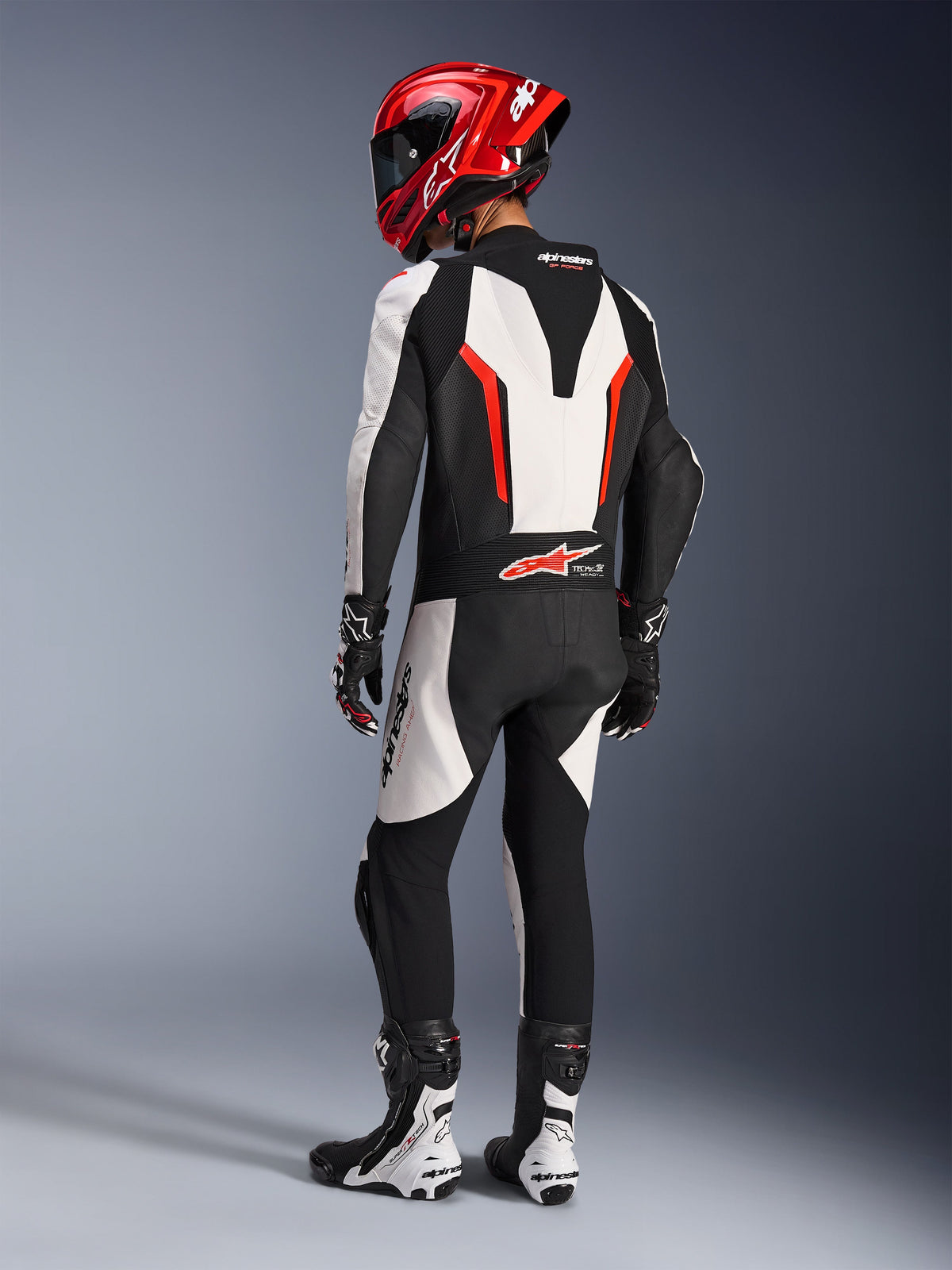Alpinestars GP Force V2 Lederkombi – 1-teilig, Weiß Schwarz Rot Fluo, Rückansicht eines Fahrers in voller Rennausrüstung, einschließlich rotem Helm, schwarz-weißen Stiefeln und Handschuhen, hebt den aerodynamischen Rückenhöcker und die ergonomischen Stretch-Paneele hervor.