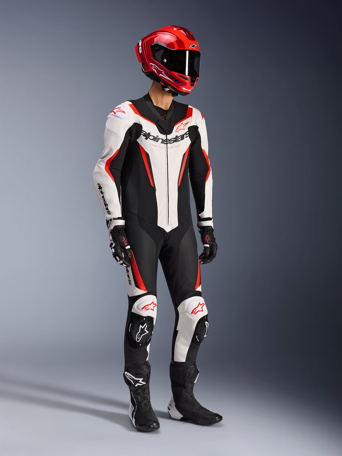 Alpinestars GP Force V2 Lederkombi 1-teilig in Weiß Schwarz und Rot Fluo, Ganzkörper-Studioaufnahme eines Fahrers in kompletter Rennausrüstung mit rotem Helm, schwarzen Handschuhen und schwarzen Stiefeln, aerodynamische Rindslederkonstruktion mit markantem Branding und integrierten Knieschleifern.
