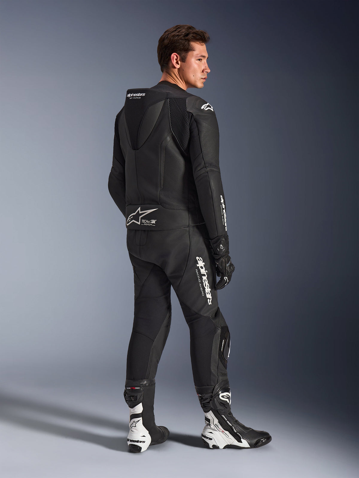 Male model wearing Alpinestars GP Force V2 Leather Suit 2-Piece in Black with white logos, rear three-quarter view showing the aerodynamic speed hump and Tech-Air Ready branding, paired with black and white racing boots and gloves, professional sport and track motorcycle gear.