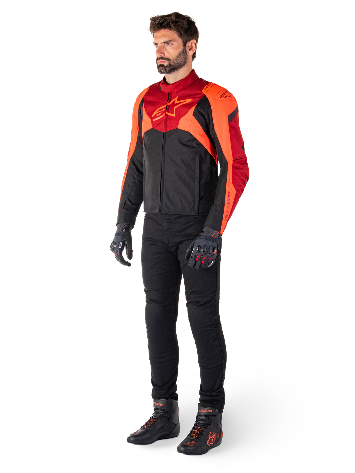 Männlicher Fahrer in der Alpinestars T-Jaws V4 Wasserdichten Jacke in Schwarz Rot Fluo Dunkelrot, kombiniert mit schwarzer Motorradhose, Handschuhen und Motorradstiefeln, sportlich geschnittenes Textildesign mit markantem Branding auf der Brust und einer aggressiven Silhouette.