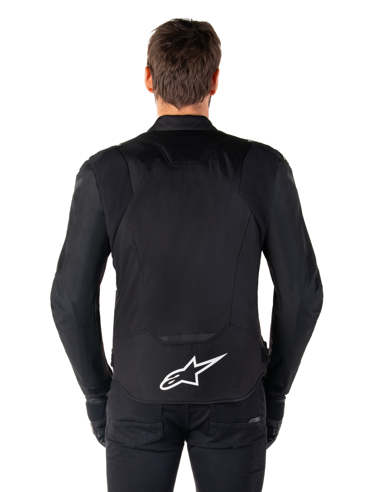 Alpinestars T-Jaws V4 Waterproof Jacke, schwarze Motorradjacke in der Rückansicht, mit weißem Alpinestars-Logo auf dem unteren Rücken, verlängerte Rückenpartie für optimale Abdeckung, getragen von Modell mit schwarzer Hose und Handschuhen, sportliches Textildesign