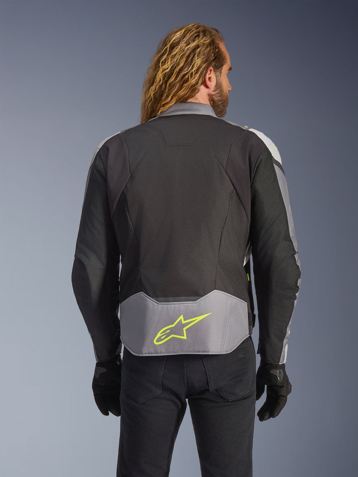 Alpinestars T-Jaws V4 Wasserdichte Jacke, Schwarz Eisgrau Dunkelgrau, Rückansicht am Model, mit einem hellgrauen Einsatz am unteren Rücken und einem markanten neongelben Stern-Logo, mehrlagige Textilkonstruktion, sportliche Silhouette, getragen mit schwarzen Handschuhen und einer dunklen Hose.