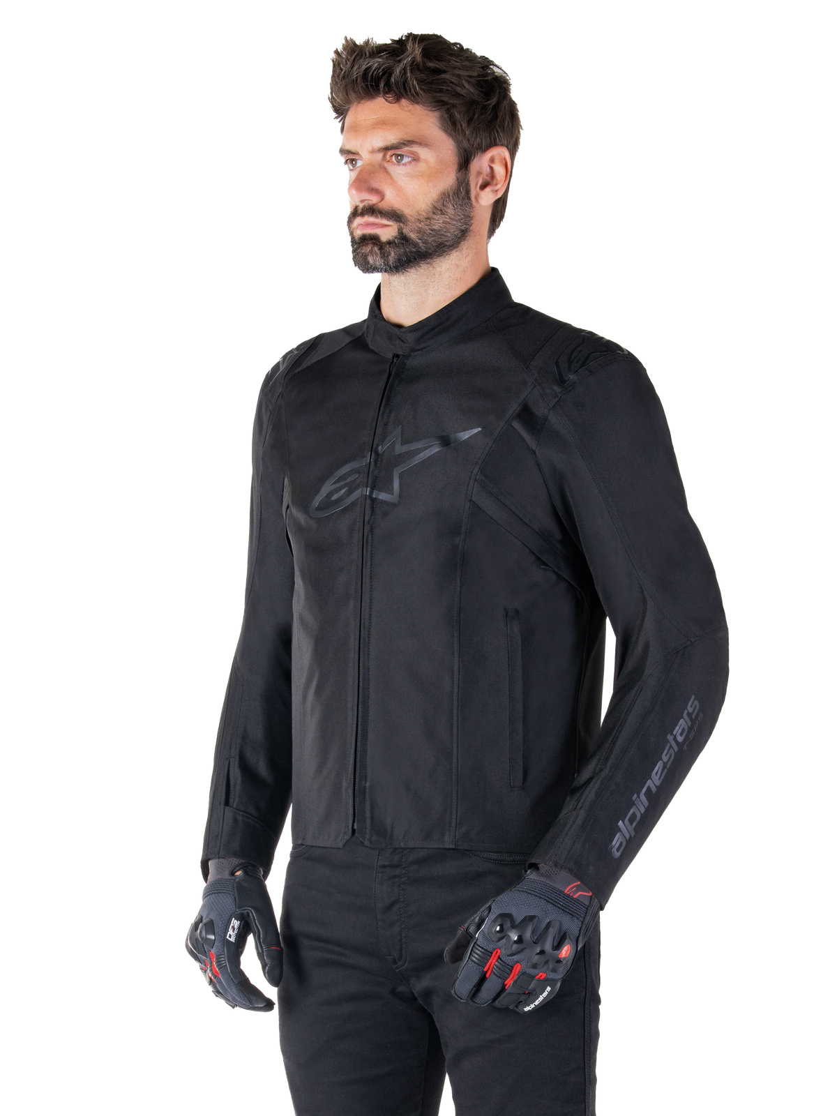 Model trägt Alpinestars T-SPS V2 Wasserdichte Jacke, Schwarz, kombiniert mit einer schwarzen Hose und grauen Motorradhandschuhen mit roten Akzenten, mit dezentem schwarzem Brustlogo und Branding am Ärmel, Textiljacke im Sportschnitt für den Straßeneinsatz.