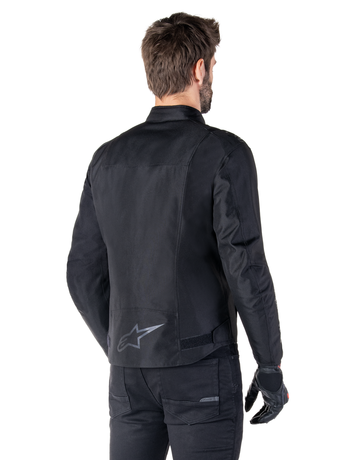 Alpinestars T-SPS V2 wasserdichte Jacke, Schwarz, Rückansicht an einem männlichen Model, mit reflektierendem Stern-Logo am unteren Rücken, verlängertem Rückenprofil und Taillenverstellriemen, kombiniert mit schwarzer Motorradhose und Handschuhen