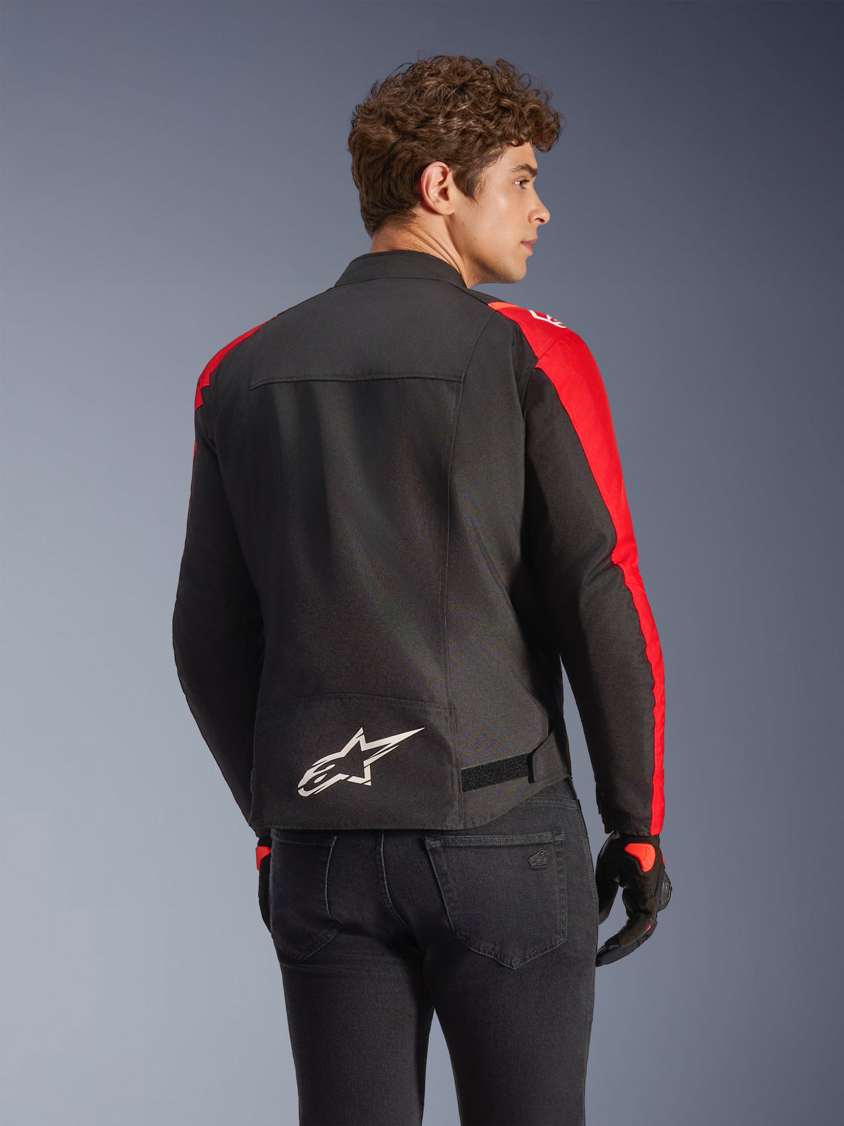 Alpinestars T-SPS V2 Waterproof Jacket in Black and Red, back view on a model, featuring a white Alpinestars logo on the lower back, vibrant red sleeve panels, and a sport-cut silhouette, worn with black gloves and jeans.