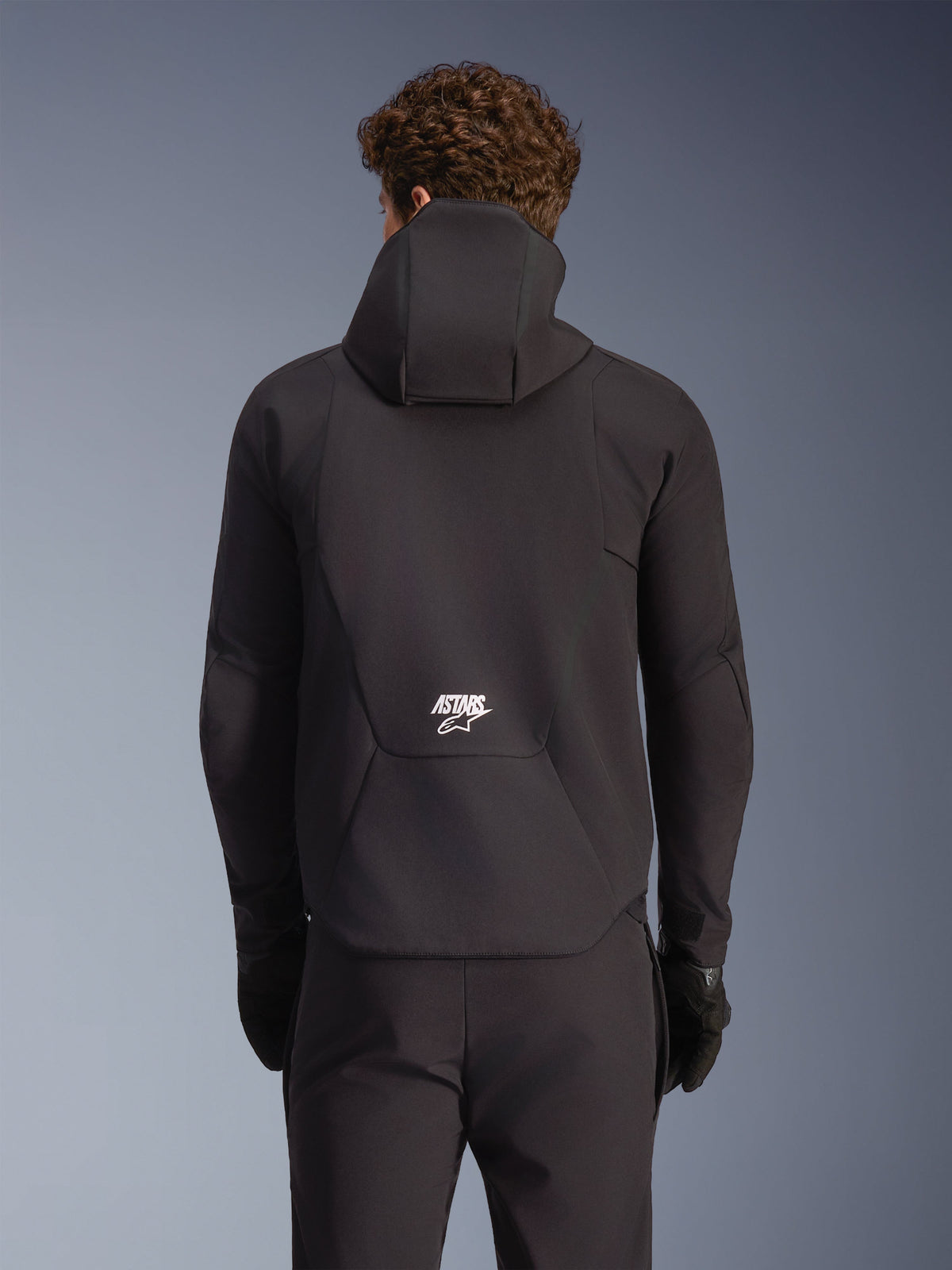 Alpinestars Aeroshell Wasserdichte Jacke, schwarze Motorrad-Softshell-Jacke, Rückansicht am Modell mit Kapuze und weißem Logo.