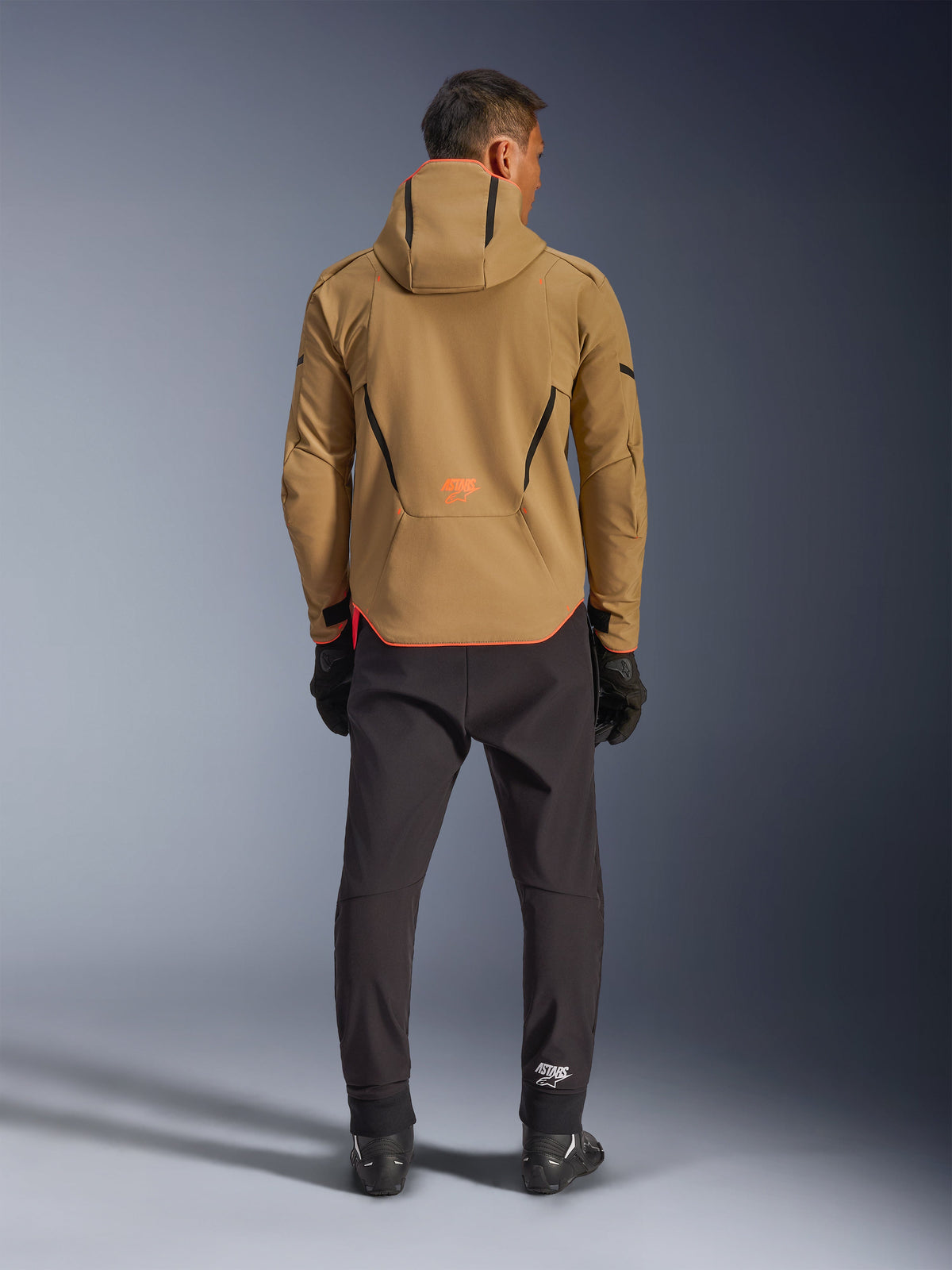 Alpinestars Aeroshell Waterproof Jacket in Dark Khaki Black Orange Fluo, Rückansicht eines Mannes, der die Softshell-Jacke mit Kapuze trägt, mit schwarzen Paspeln und orangefarbenem Logo, kombiniert mit schwarzer technischer Hose und Handschuhen.