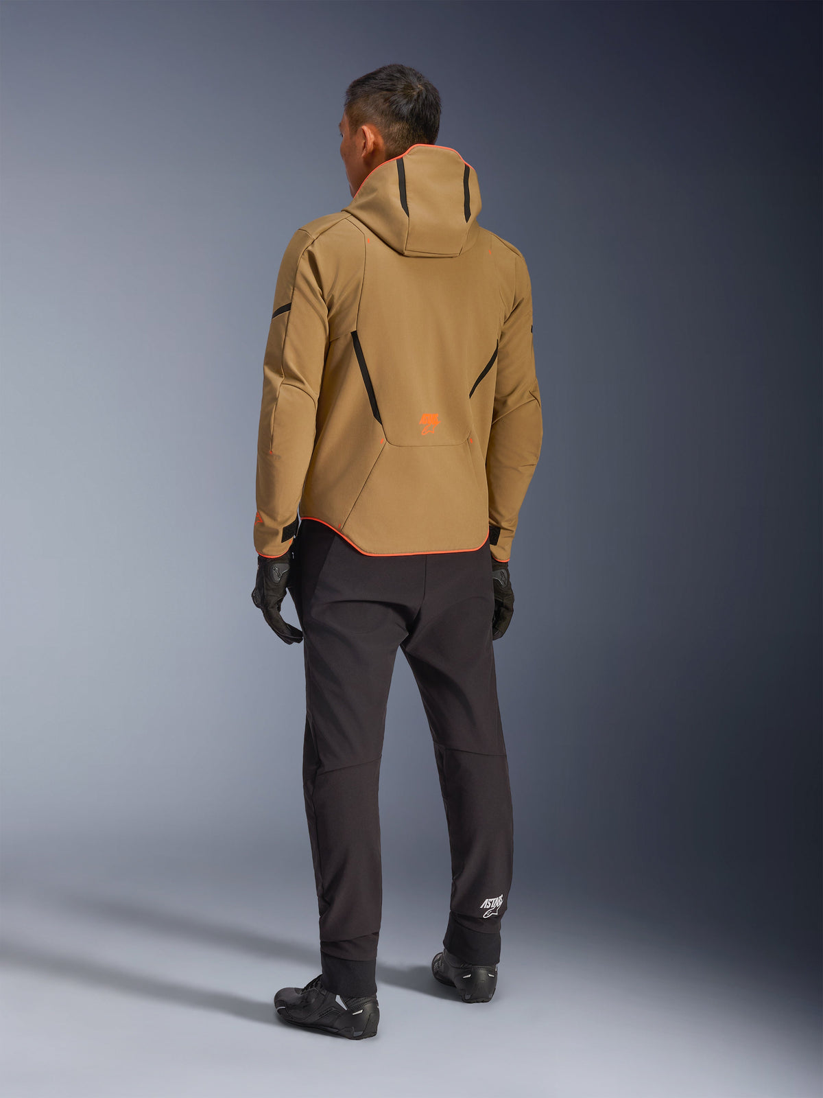 Alpinestars Aeroshell Wasserdichte Jacke, Dark Khaki Black Orange Fluo, Rückansicht eines Fahrers, der die Softshell-Jacke mit Kapuze trägt, kombiniert mit schwarzer Hose und Handschuhen, mit schwarzen geometrischen Akzenten und einem orangefarbenen Logo am unteren Rücken.