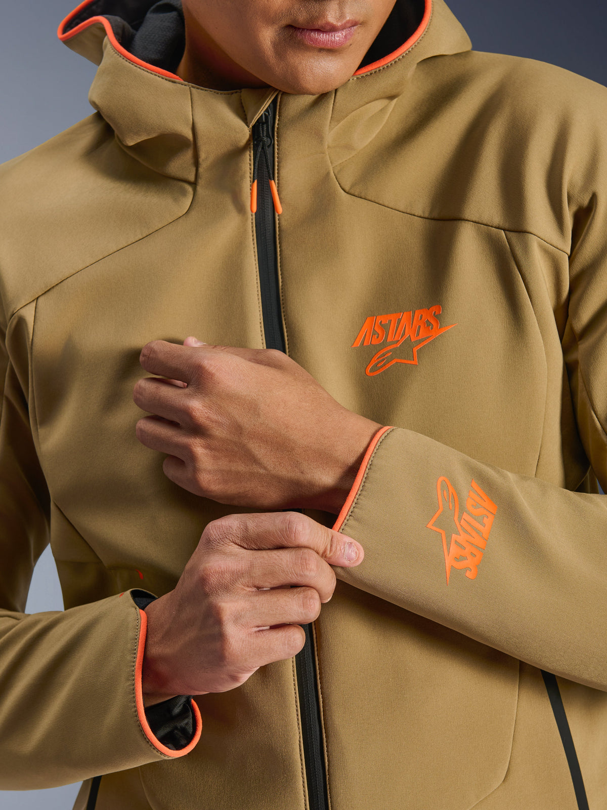 Alpinestars Aeroshell Wasserdichte Jacke, Dunkles Khaki und Orange Fluo, Nahaufnahme der Vorderseite und des Ärmels mit orangefarbenen ASTARS-Logos, orangefarbene Paspeln an Kapuze und Ärmelbündchen, schwarzer wasserdichter Reißverschluss.