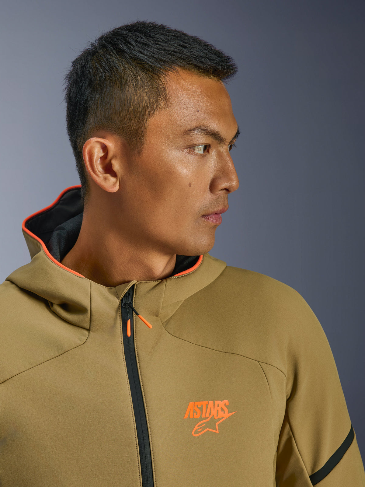 Alpinestars Aeroshell WP Jacke, Dark Khaki und Orange Fluo, Nahaufnahme im Seitenprofil, mit orangefarbenem ASTARS-Logo auf der Brust und orange gefütterter Kapuze, moderner Crossover-Softshell-Hoodie zum Motorradfahren