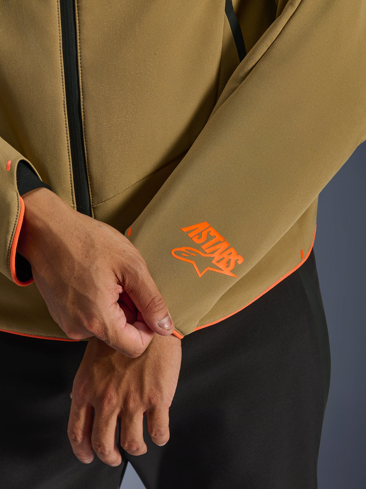 Alpinestars Aeroshell Waterproof Jacke, Dunkelkhaki mit Orange Fluo Akzenten, Nahaufnahme des Ärmelbündchens und des unteren Saums mit leuchtend orangefarbenem Astars-Logo, neon-orangener Paspelierung, wasserdichter Frontreißverschluss, sportliche Softshell-Konstruktion