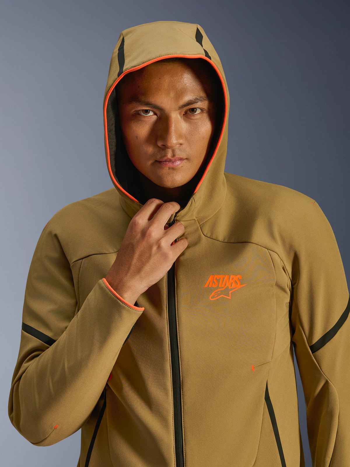 Alpinestars Aeroshell wasserdichte Jacke, Dark Khaki Black Orange Fluo, Modell trägt die Jacke mit aufgesetzter Kapuze, mit leuchtend orangefarbenen Fluo-Besätzen und einem orangefarbenen ASTARS-Logo, elegante schwarze Akzente, moderne wasserdichte Sport-Softshell