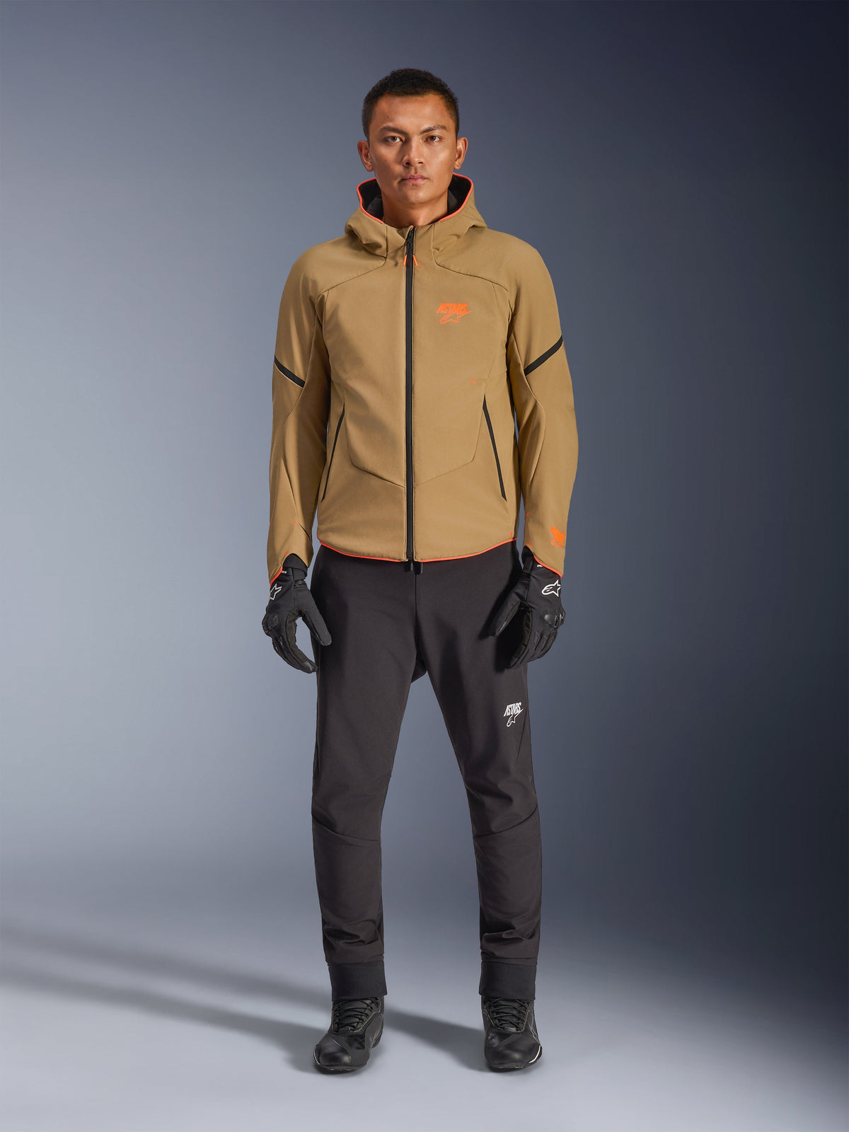 Alpinestars Aeroshell Waterproof Jacke, männliches Model in Dark Khaki mit schwarzen und neon-orangen Akzenten, orangenes Logo auf der Brust und Kapuzendesign, kombiniert mit schwarzer Hose und schwarzen Motorradhandschuhen, lässiger Sport-Touring-Stil