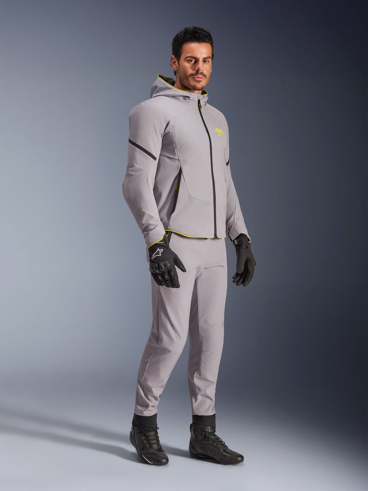 Alpinestars Aeroshell Waterproof Jacket, Mid Gray and Yellow Fluo, worn by model with matching gray pants, black riding gloves, and black boots, modern hoodie-style motorcycle gear for casual and sport riding.